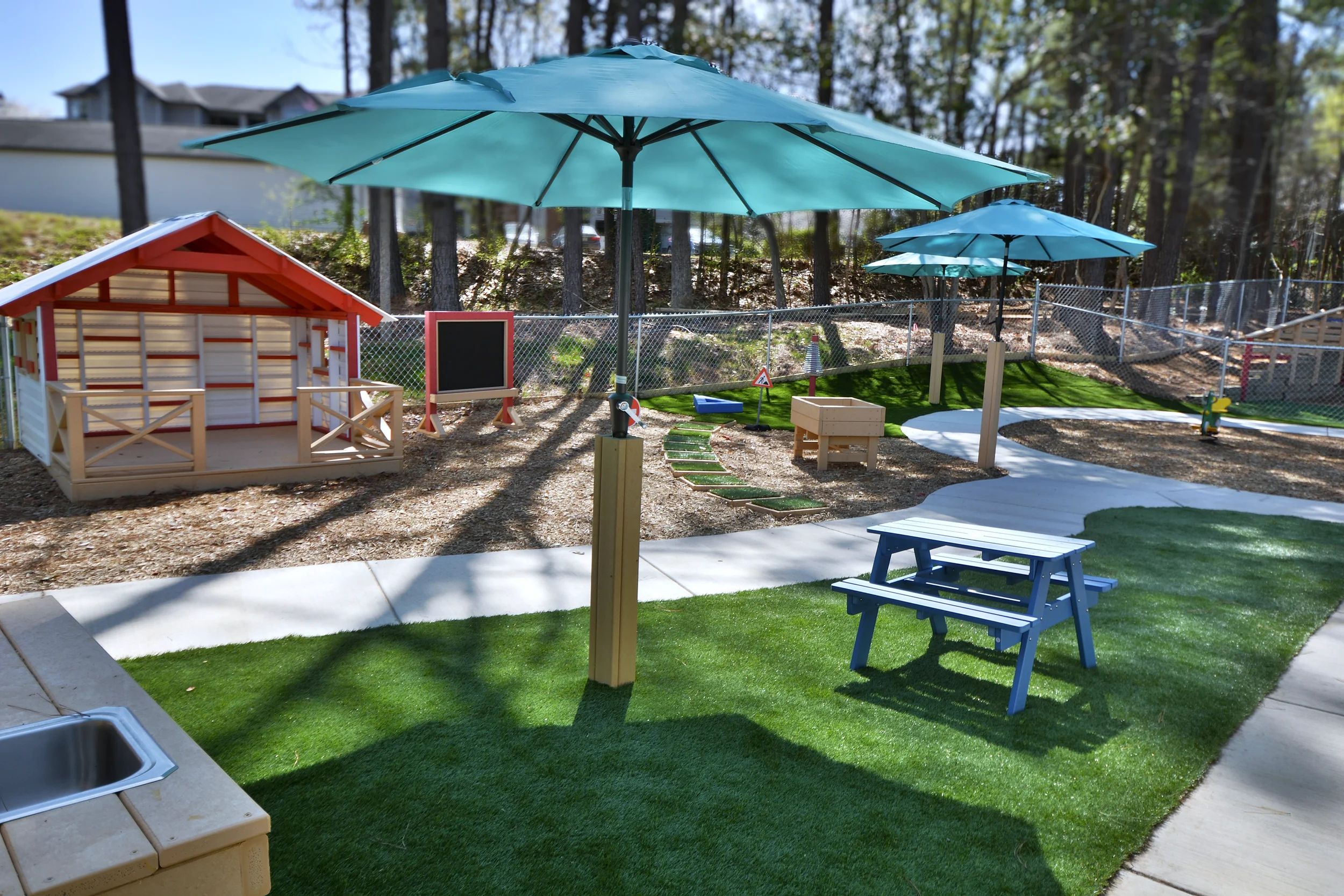 Children's outdoor play area with large blue umbrellas, a small red and white playhouse, a sandbox, a picnic table, and artificial turf, fenced with trees in the background.