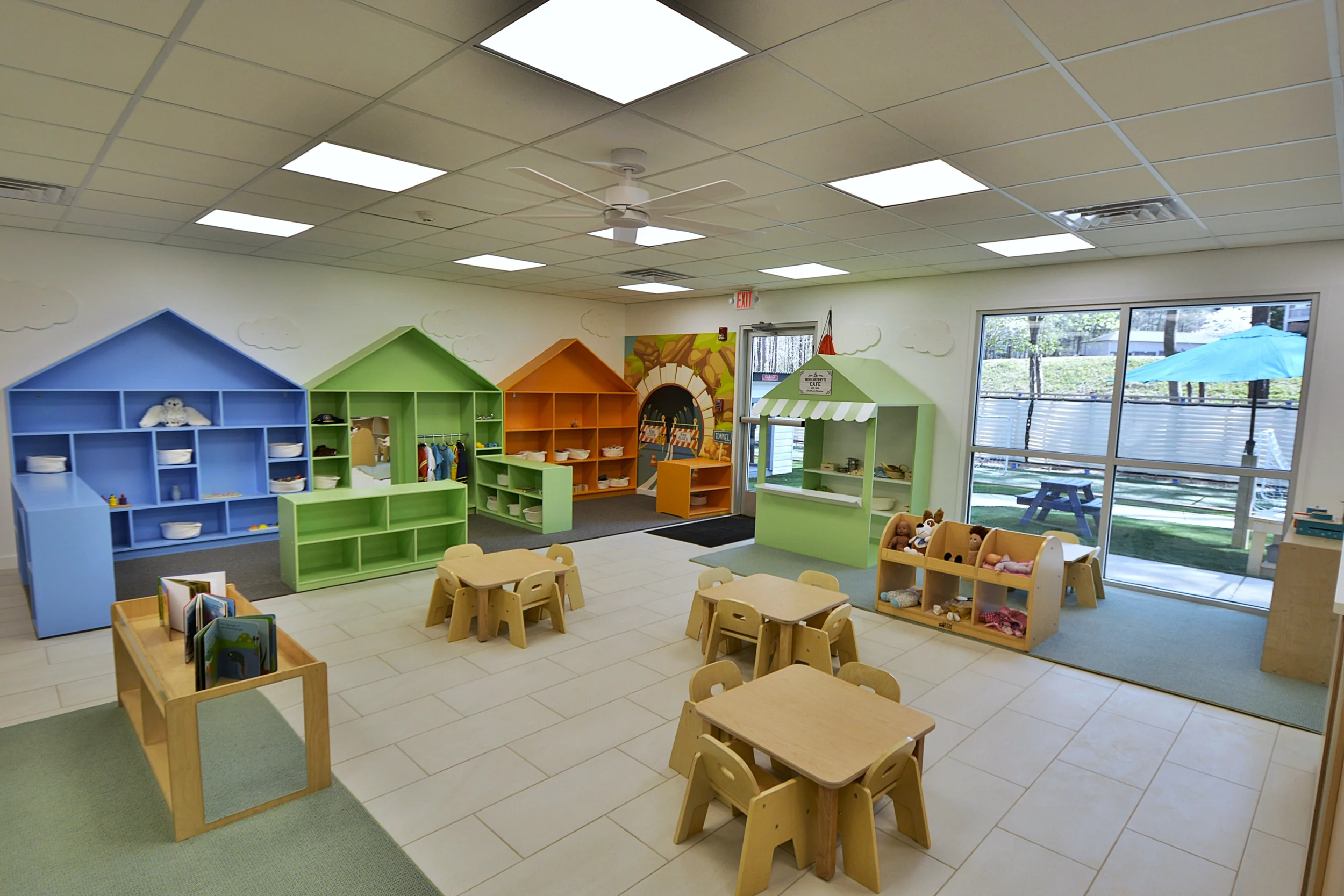Interior of a children's classroom with colorful house-shaped shelves, small tables and chairs, stuffed toys, and outdoor view through large windows.