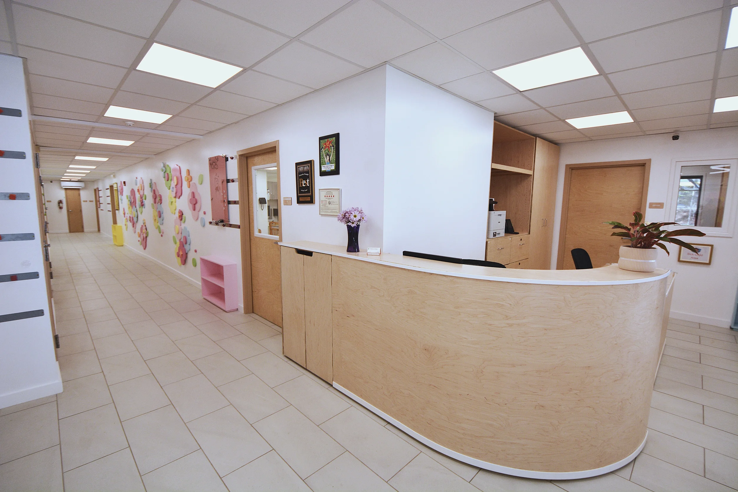 Reception area with a curved wooden counter, flowers in a purple vase, and framed pictures on the wall, with a hallway decorated with colorful wall art.