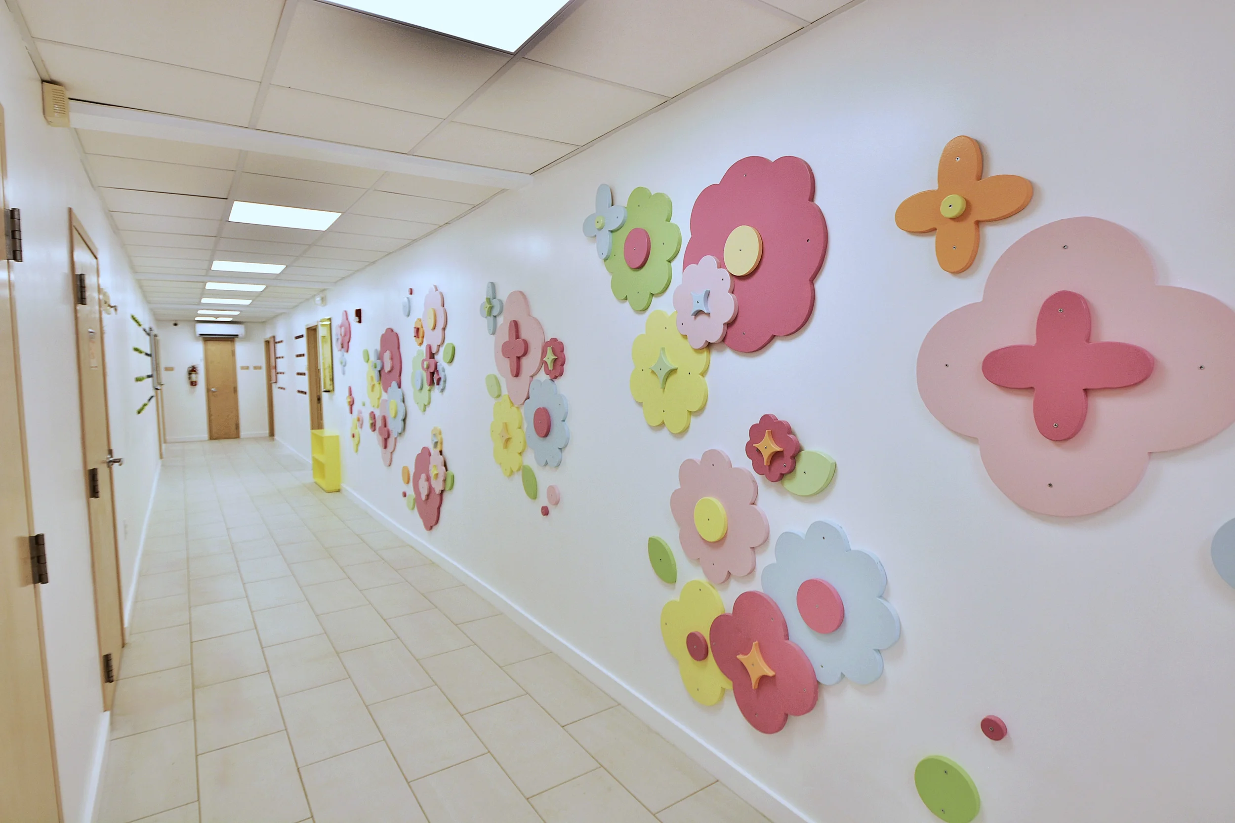 A hallway with a white wall decorated with large, colorful, floral-shaped wall art in pastel shades of pink, green, yellow, orange, and white. The hallway has a tiled floor, closed doors on the left, and a drop ceiling with fluorescent lighting.