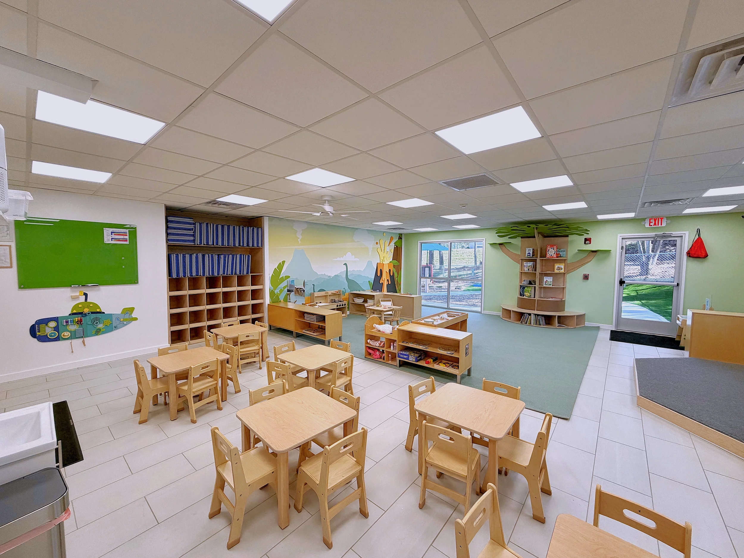 A brightly lit classroom with wooden tables and chairs, a green wall with a forest mural, wooden shelves filled with books, and a window showing an outdoor play area.
