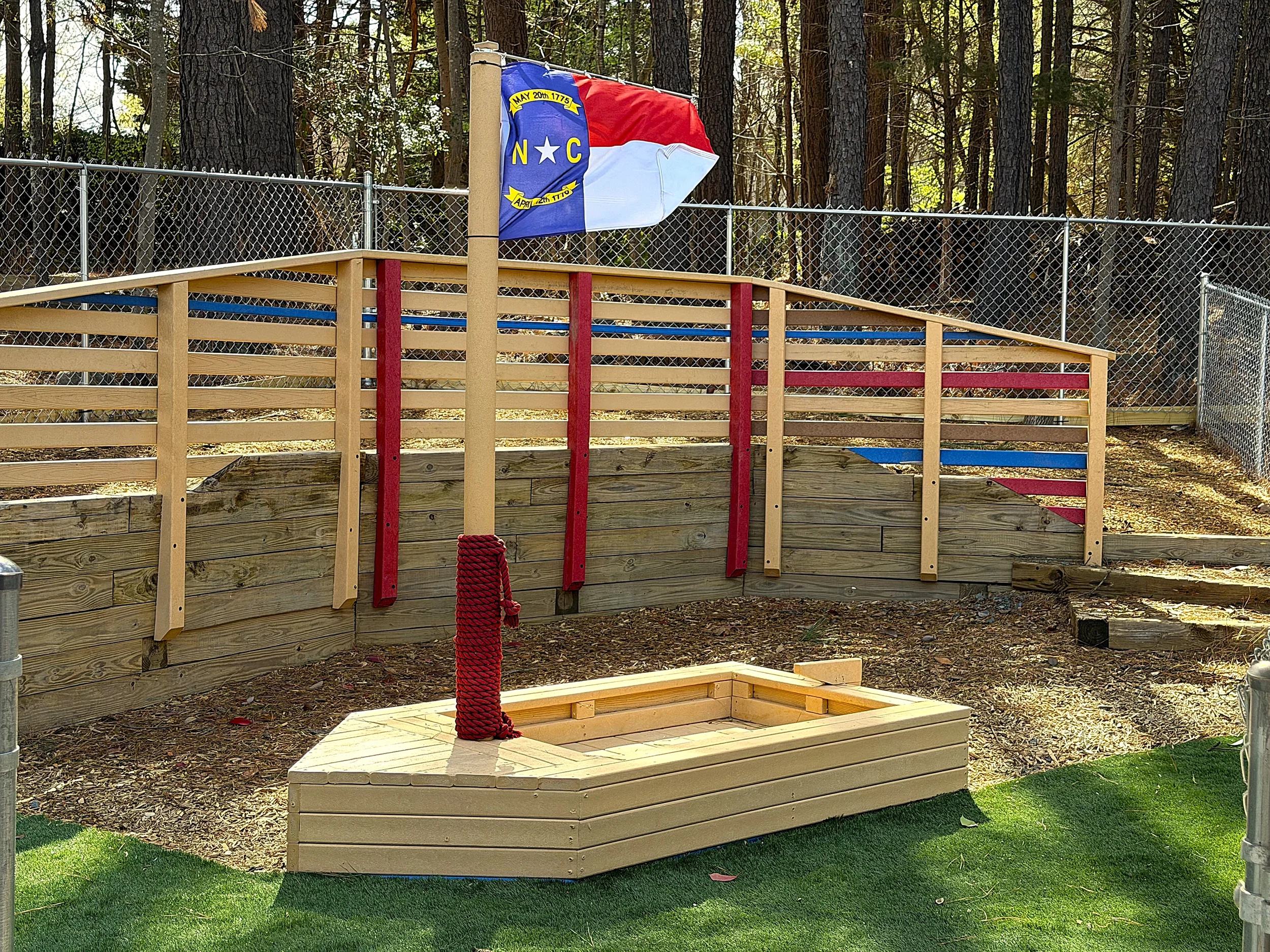 A photograph of part of the playground, featuring a North Carolina State flag that is planted into a toy wooden boat. 