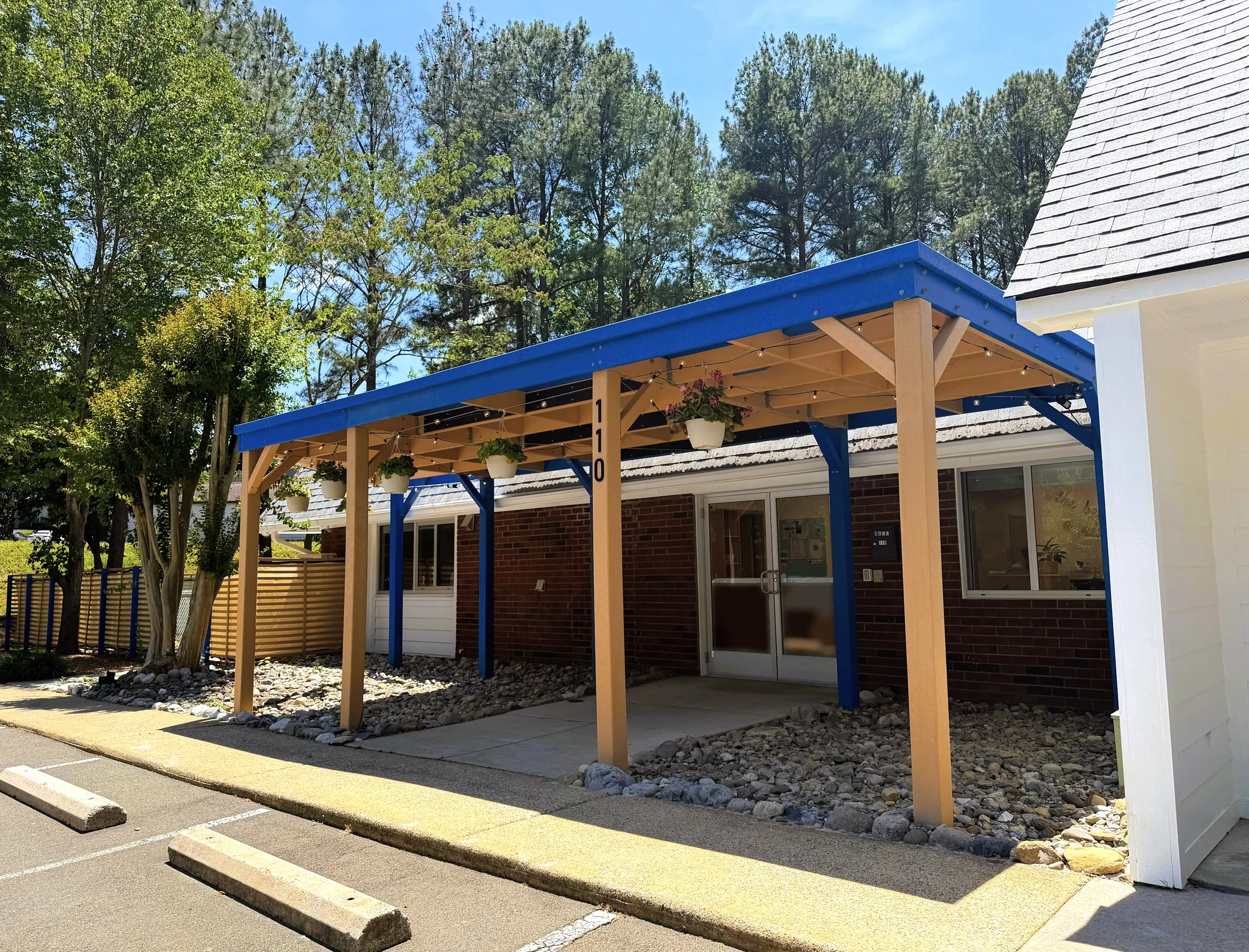 Exterior view of a building with a blue wooden canopy supported by wooden beams, decorated with hanging flower pots and string lights, surrounded by trees and a parking lot.