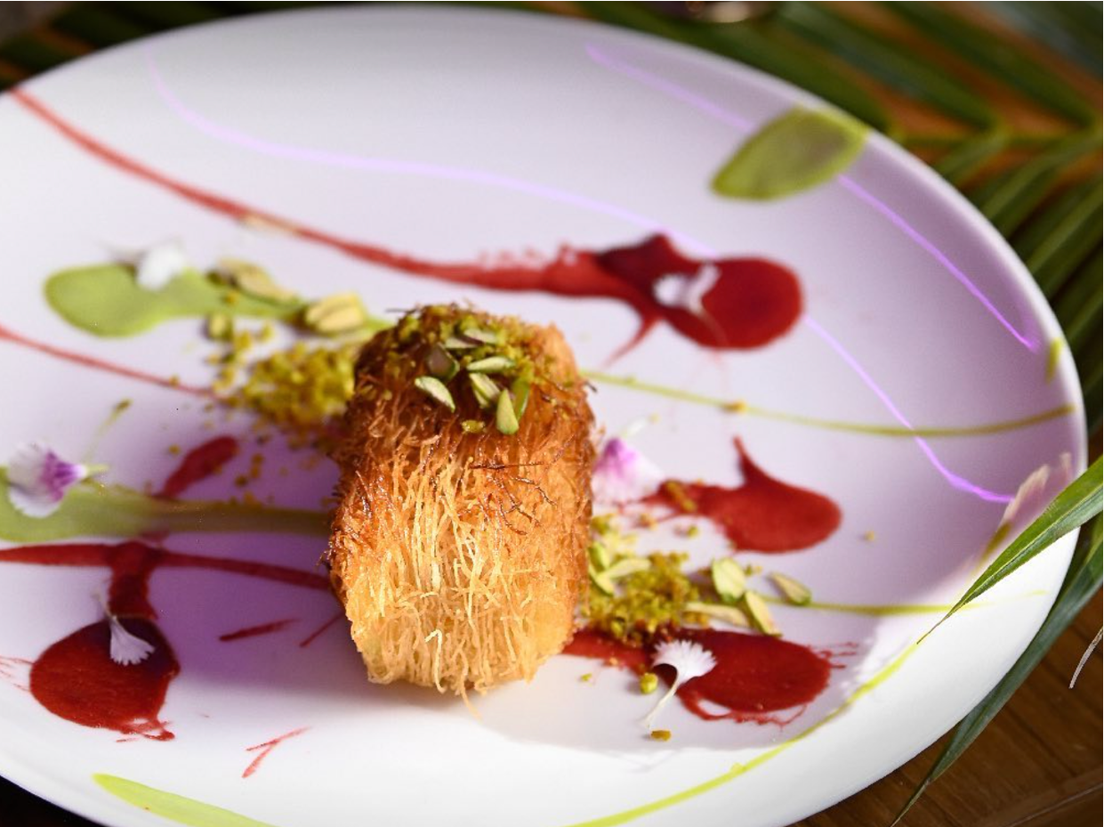 An ornate dessert with a fried, hollow, pastry cylinder topped with chopped pistachios, served on a white plate decorated with colorful sauce drizzles and edible flowers.