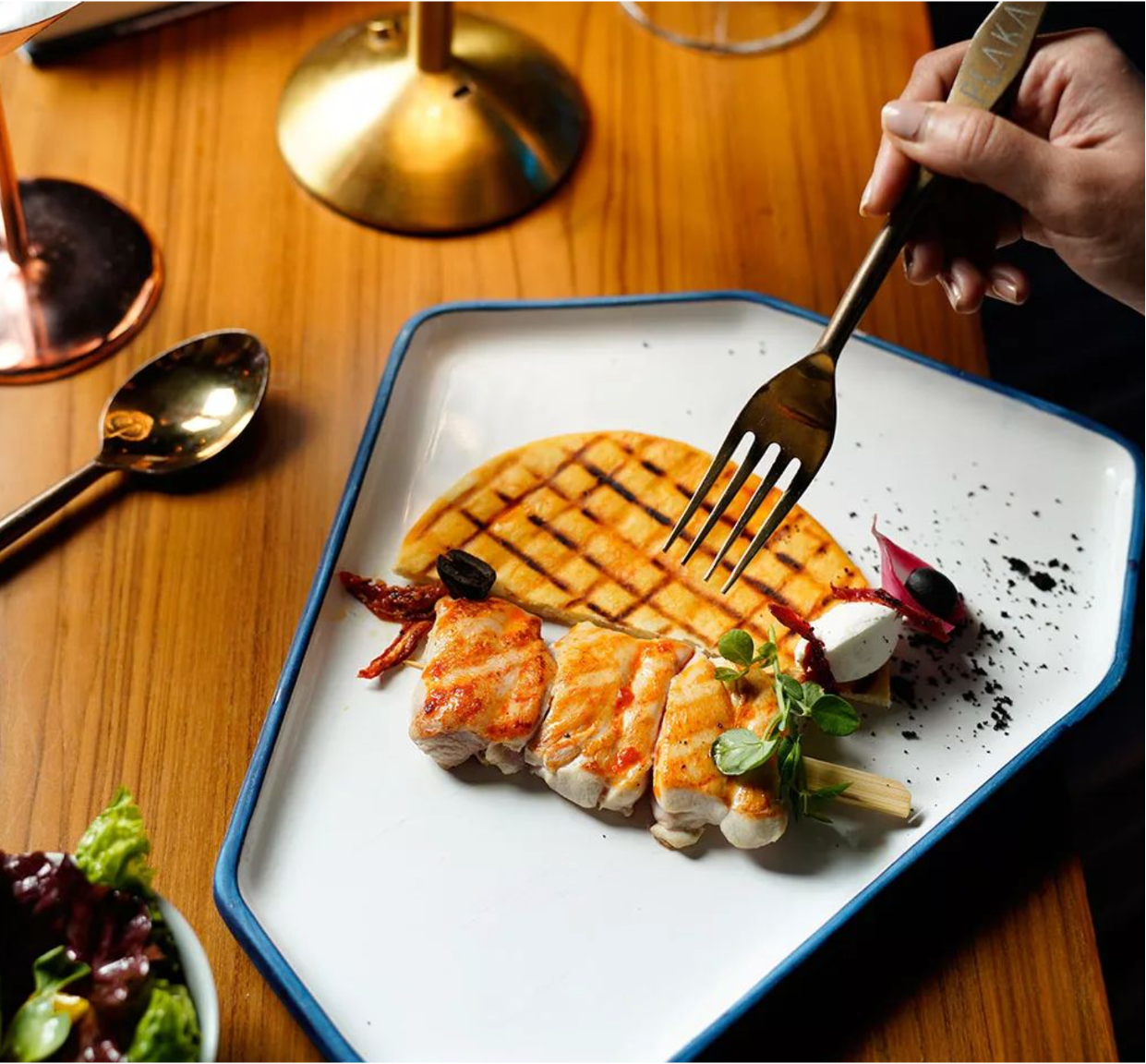 A plated meal with grilled chicken, a waffle, and garnishes on a rectangular white and blue plate. A person is holding a fork, about to eat. There are also a spoon, a copper-colored glass, a lamp, and a partially visible bowl of salad on a wooden tab