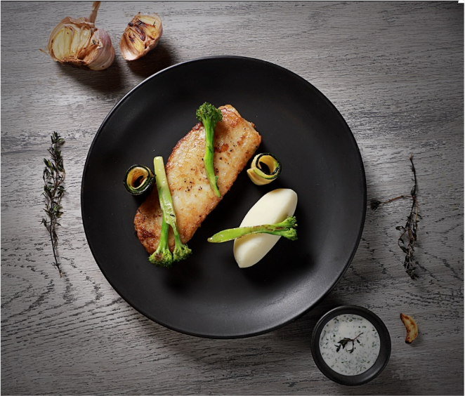 Pan-fried fish fillet with broccoli and leek ribbons, served with mashed potatoes and a herb sauce on a black plate, surrounded by garlic, thyme, and a small bowl of sauce on a gray wooden surface.