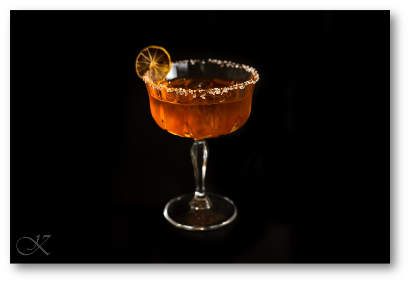 An elegant cocktail in a flared glass with a salted rim and a dried lemon garnish, set against a black background.