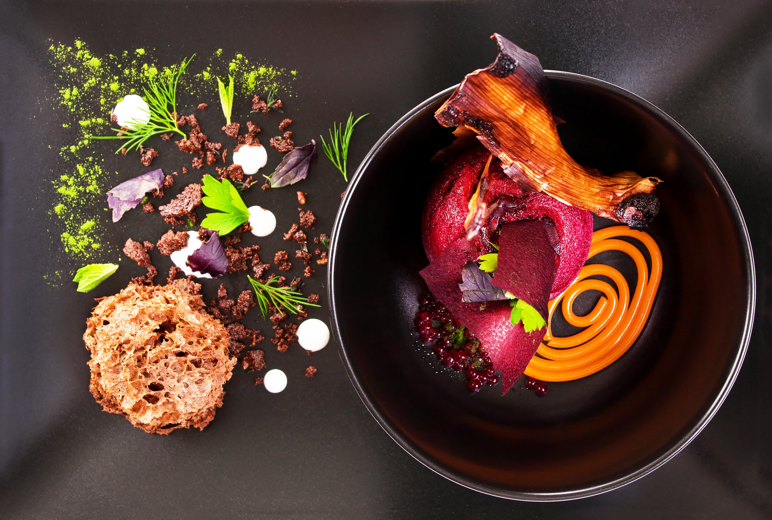 An artfully plated dessert featuring a pink sorbet or mousse topped with a caramelized sugar piece, surrounded by spiralized orange carrot ribbons, edible purple and green herbs, chocolate crumbs, and small dollops of white sauce on a dark background.