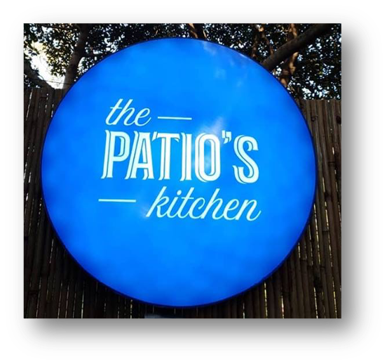 Blue circular sign with white text reading 'the patio's kitchen' outdoors against a wooden fence with trees in the background.