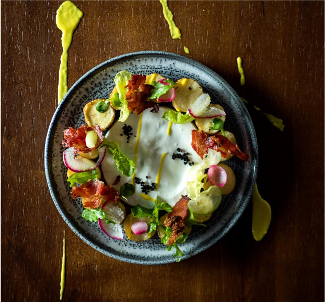 A plate of salad with bacon, radish slices, lettuce, and black sesame seeds on a dark wooden table with some yellow sauce splattered around.
