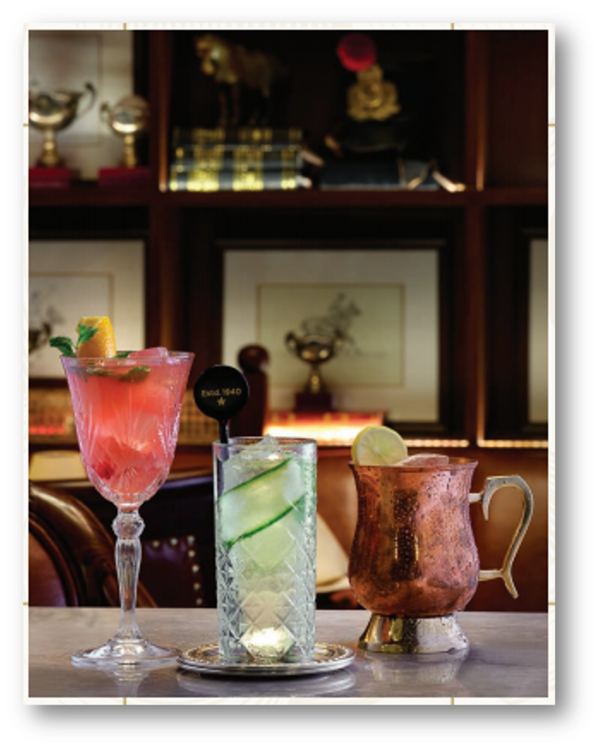 Three cocktails on a bar counter, with a wooden shelf with trophies and framed artwork in the background.