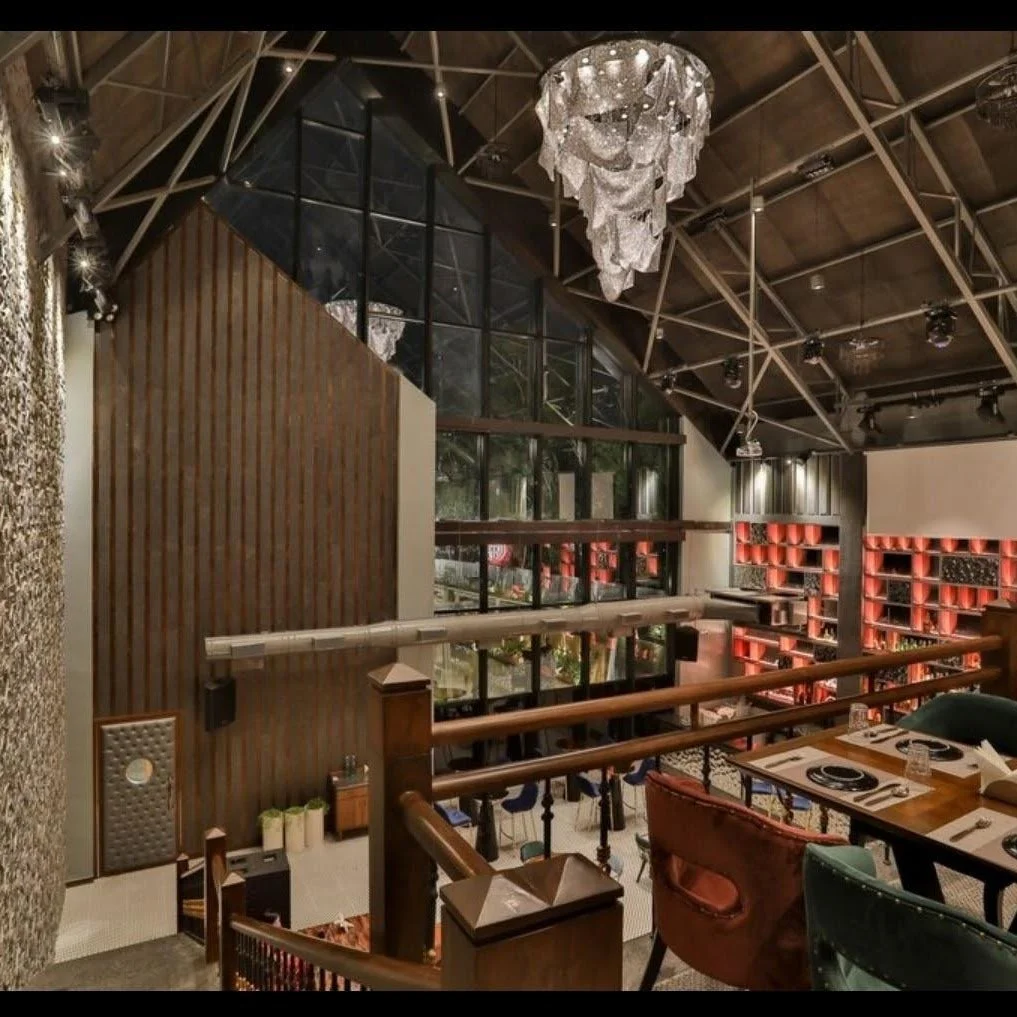 Interior view of a modern restaurant with high ceilings, large window panes, and a chandelier. There are tables set for dining with plates, glasses, and utensils. A large bookcase with red shelves decorates the far wall.