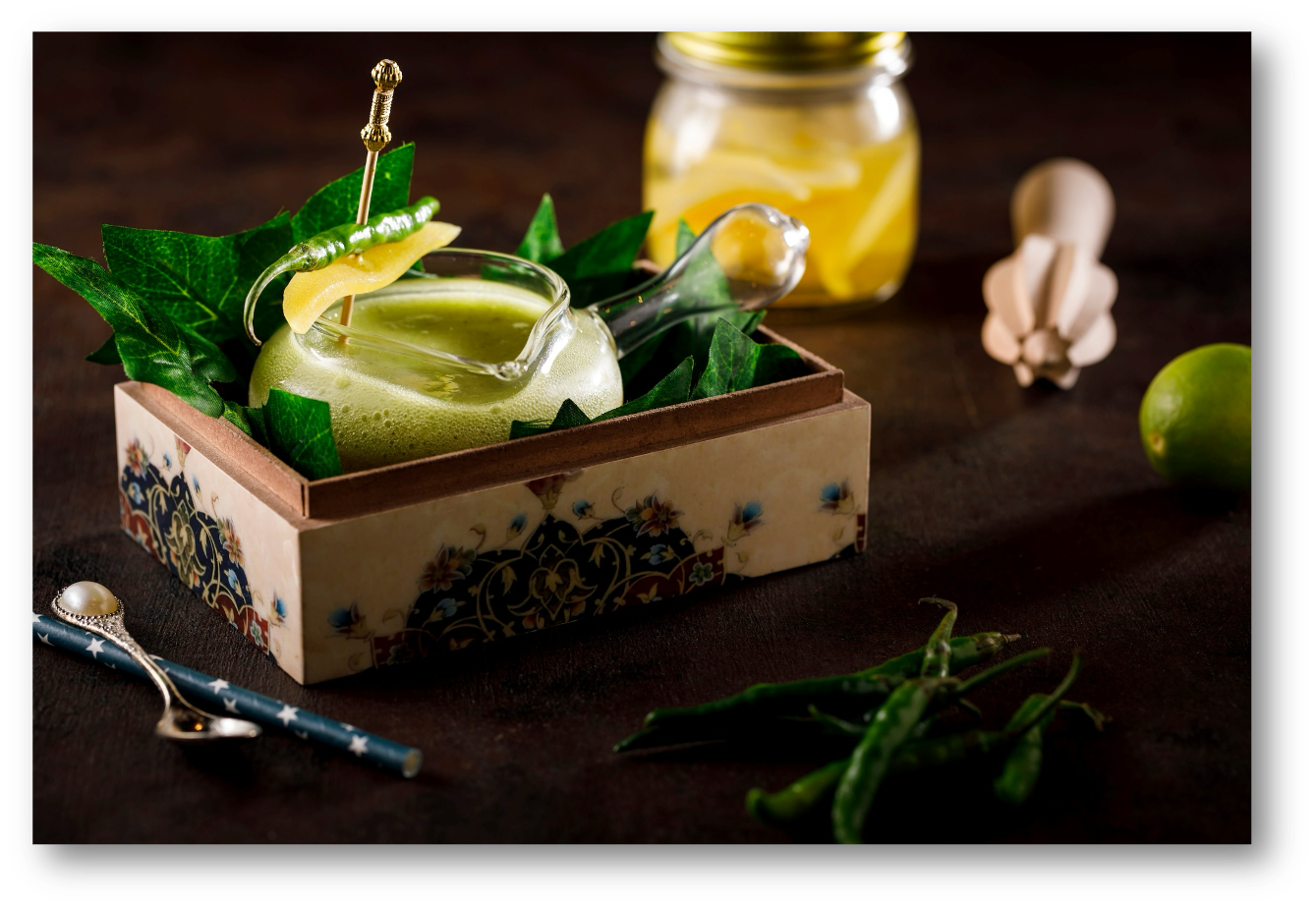 A glass teapot with a green beverage surrounded by decorative ivy leaves, a small jar of lemon slices, a lime, and a few green chili peppers on a dark surface.