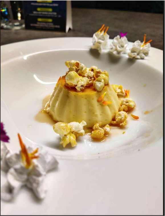 A dessert with a pudding-like base topped with caramel sauce and popcorn, garnished with edible flower petals and accompanied by whipped cream decorated with colorful edible decorations.