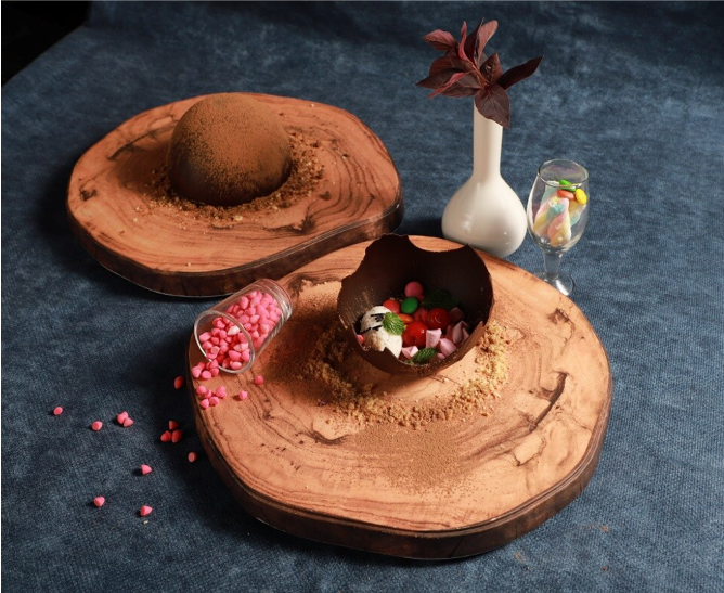 Chocolate mousse dessert on a wooden platter with colorful candies and garnishes, accompanied by a small vase with dark red leaves and a glass of candy sprinkles.