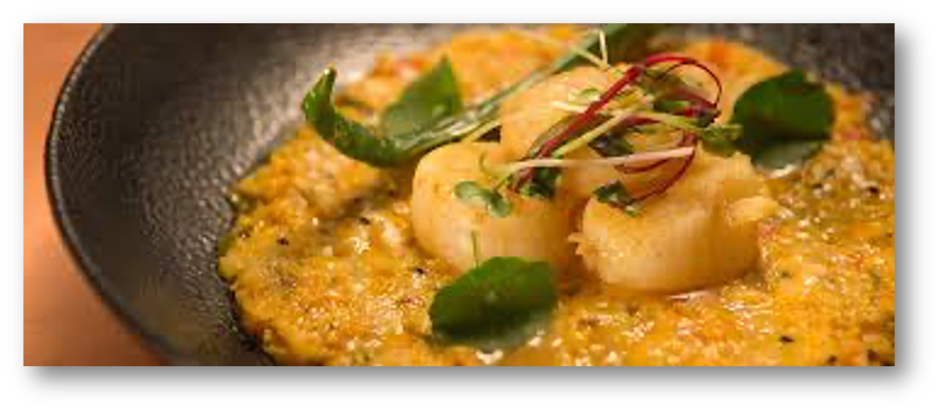 Close-up of a seafood dish with seared scallops, garnished with microgreens, served on a bed of creamy sauce in a black skillet.