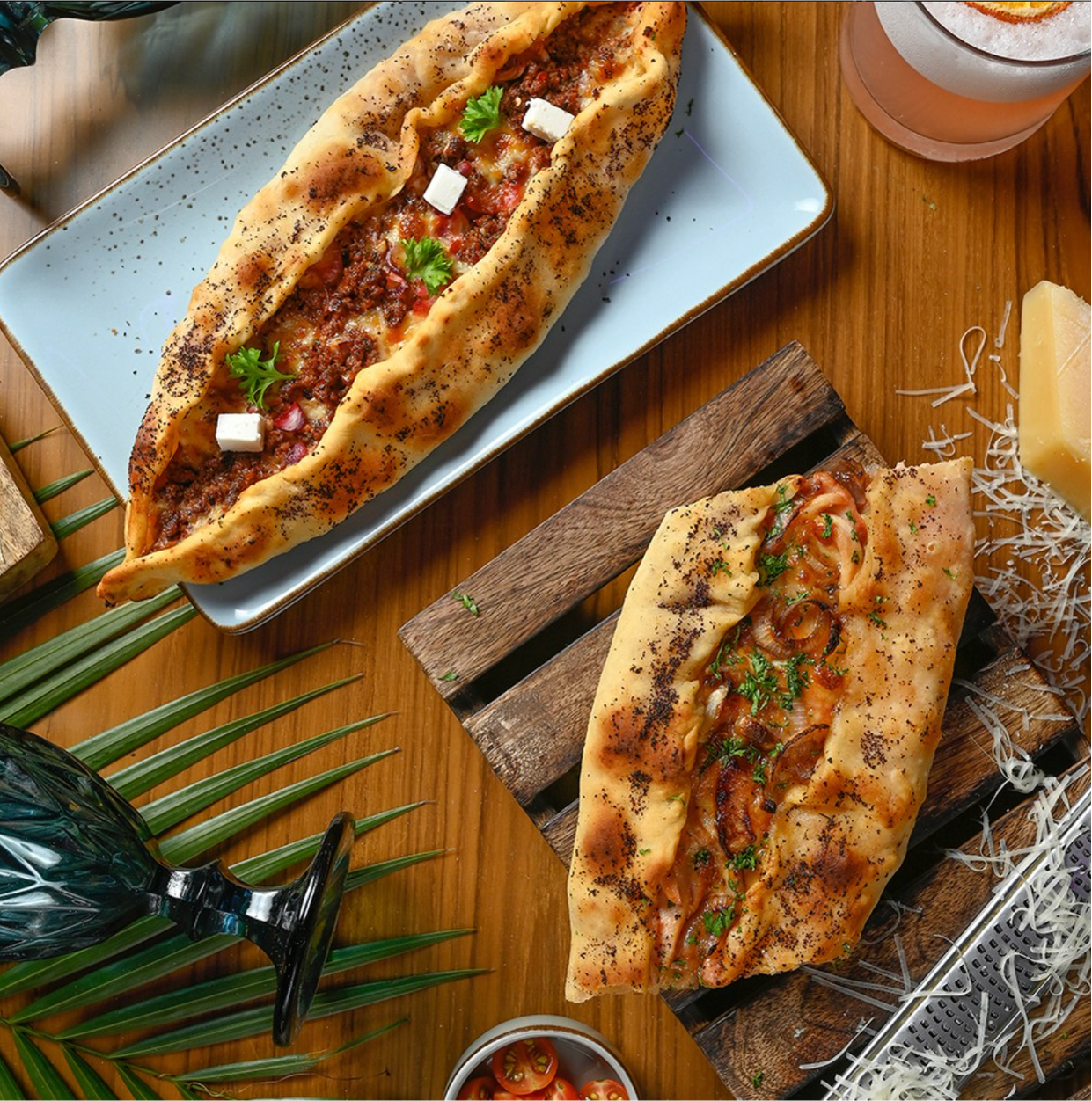 Two Turkish-style flatbreads with toppings on a wooden table. One flatbread has ground meat, cheese, tomatoes, and herbs. The other has sliced onions, tomatoes, herbs, and spices. There are also a glass of pink drink, cheese, cherry tomatoes, shredde