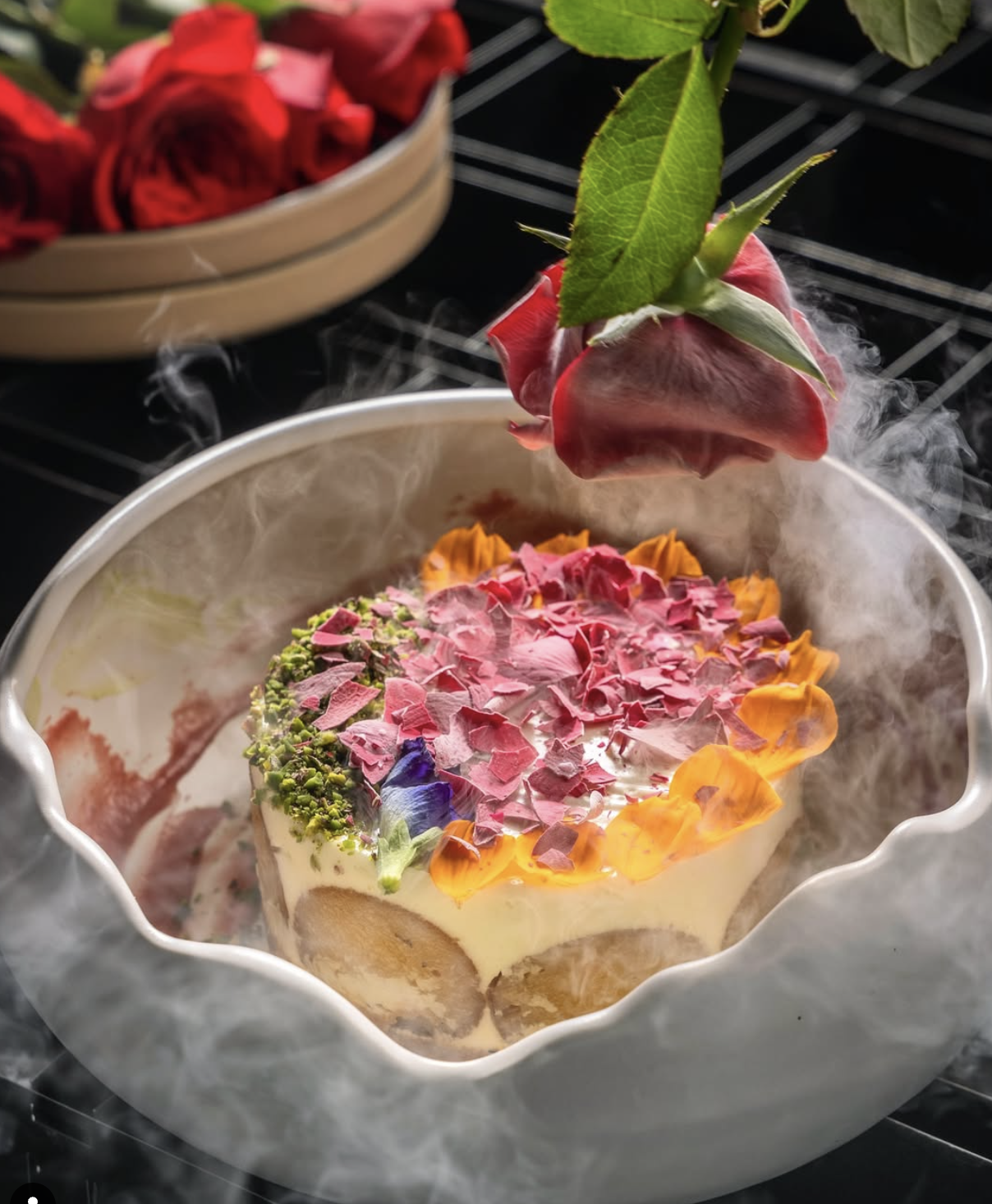 A cake decorated with colorful flower petals and greenery, surrounded by dry ice creating a misty effect.