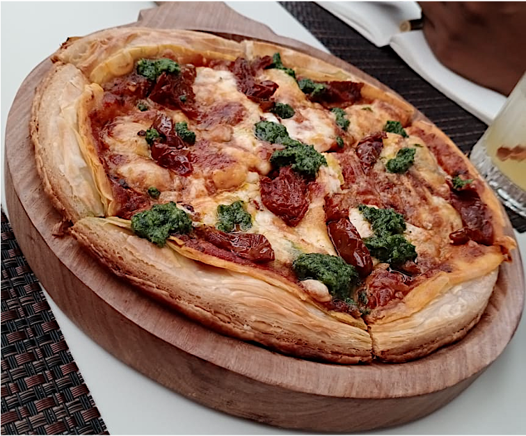 Flatbread pizza with cheese, sun-dried tomatoes, and green pesto on a wooden serving board.
