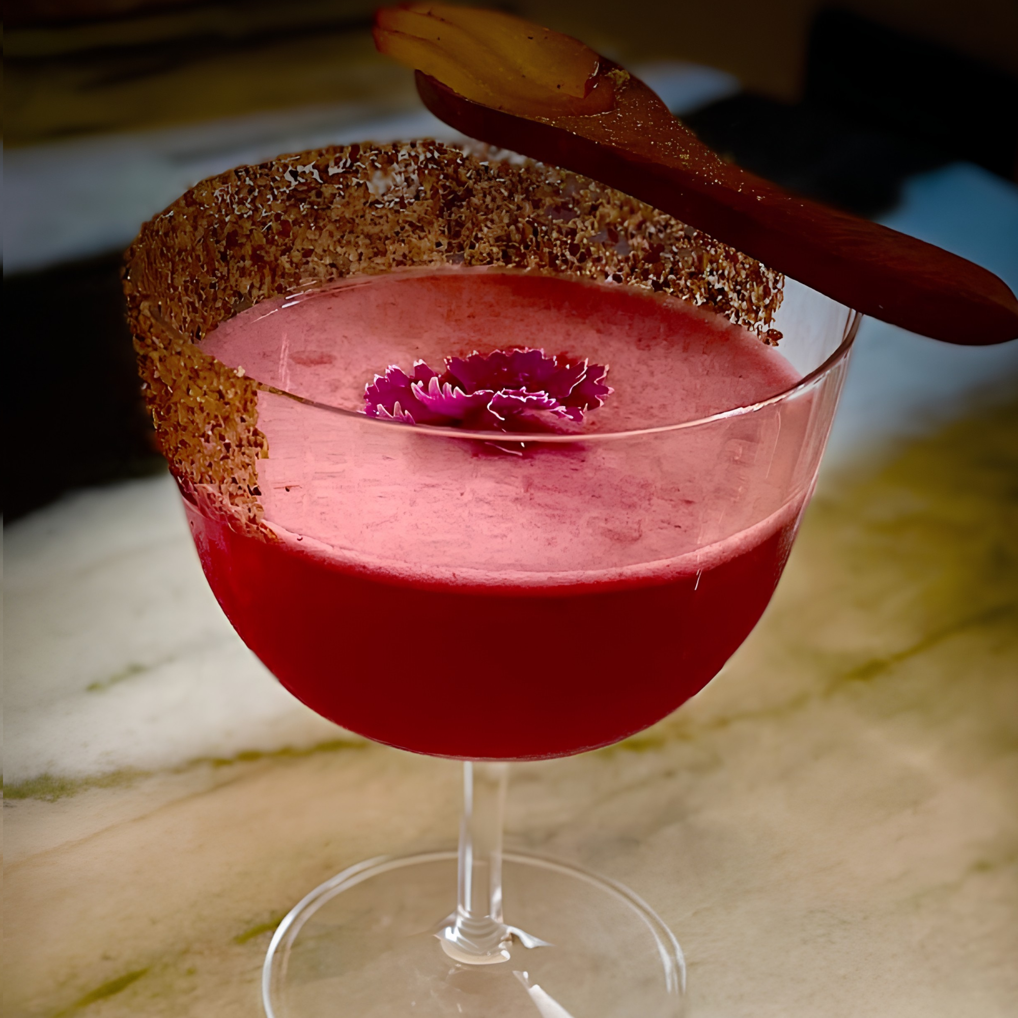 Close-up of a pink flower-shaped candle in a glass holder with a floating wax flower inside, surrounded by a red, sugar-coated candy on top, placed on a beige surface.