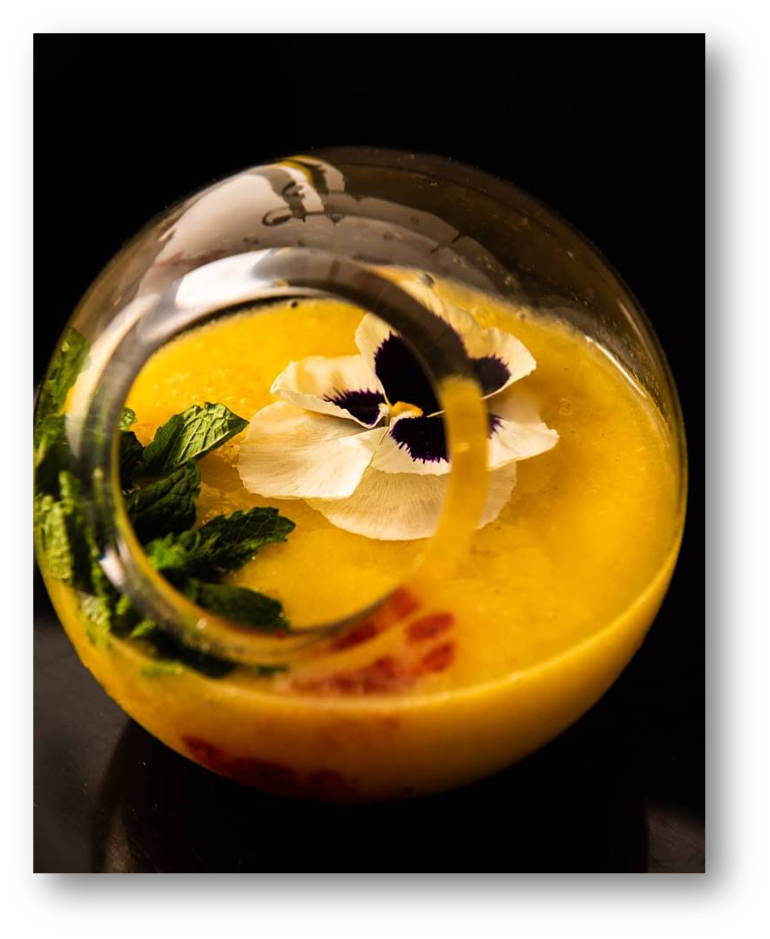 A yellow cocktail in a spherical glass garnished with a flower and mint leaves.