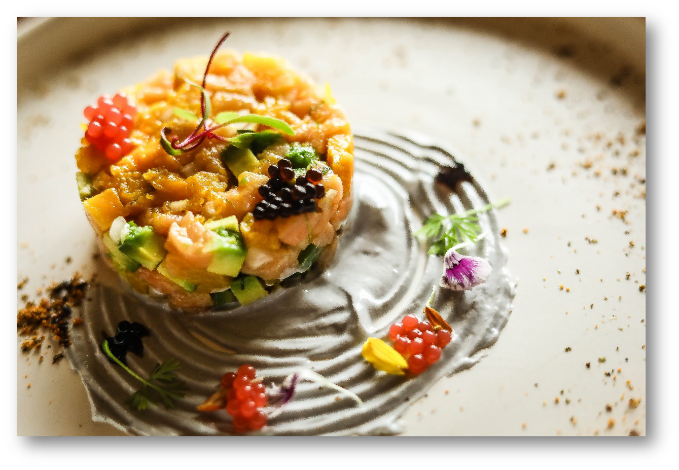 An elegantly plated dish featuring a small, colorful tart topped with diced vegetables and garnished with edible flowers and small berries, served on a white plate with artistic sauce and seasoning.