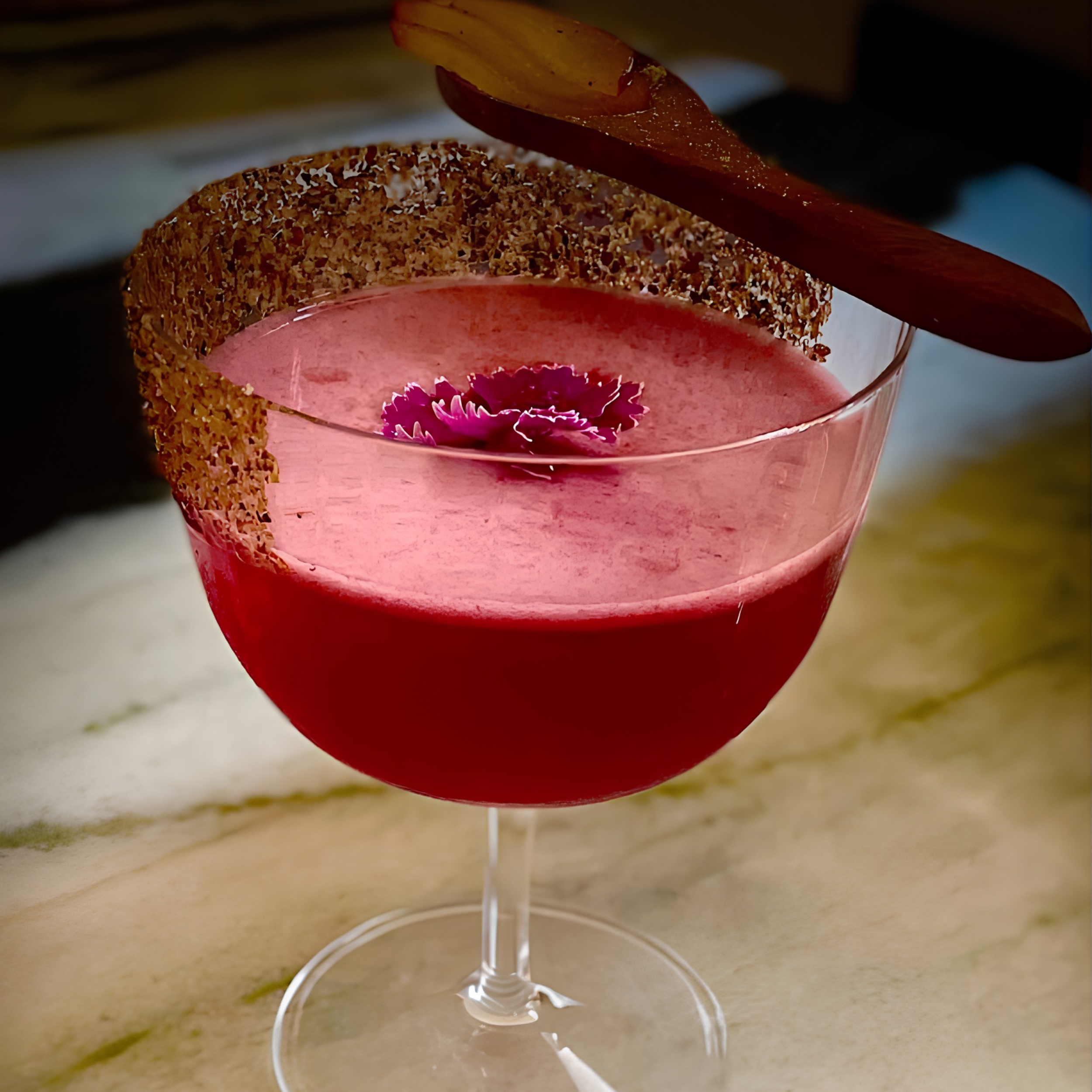 Close-up of a pink candle with a flower inside, placed in a glass holder with a wood chip border surrounding the wax, illuminated in a dim setting.