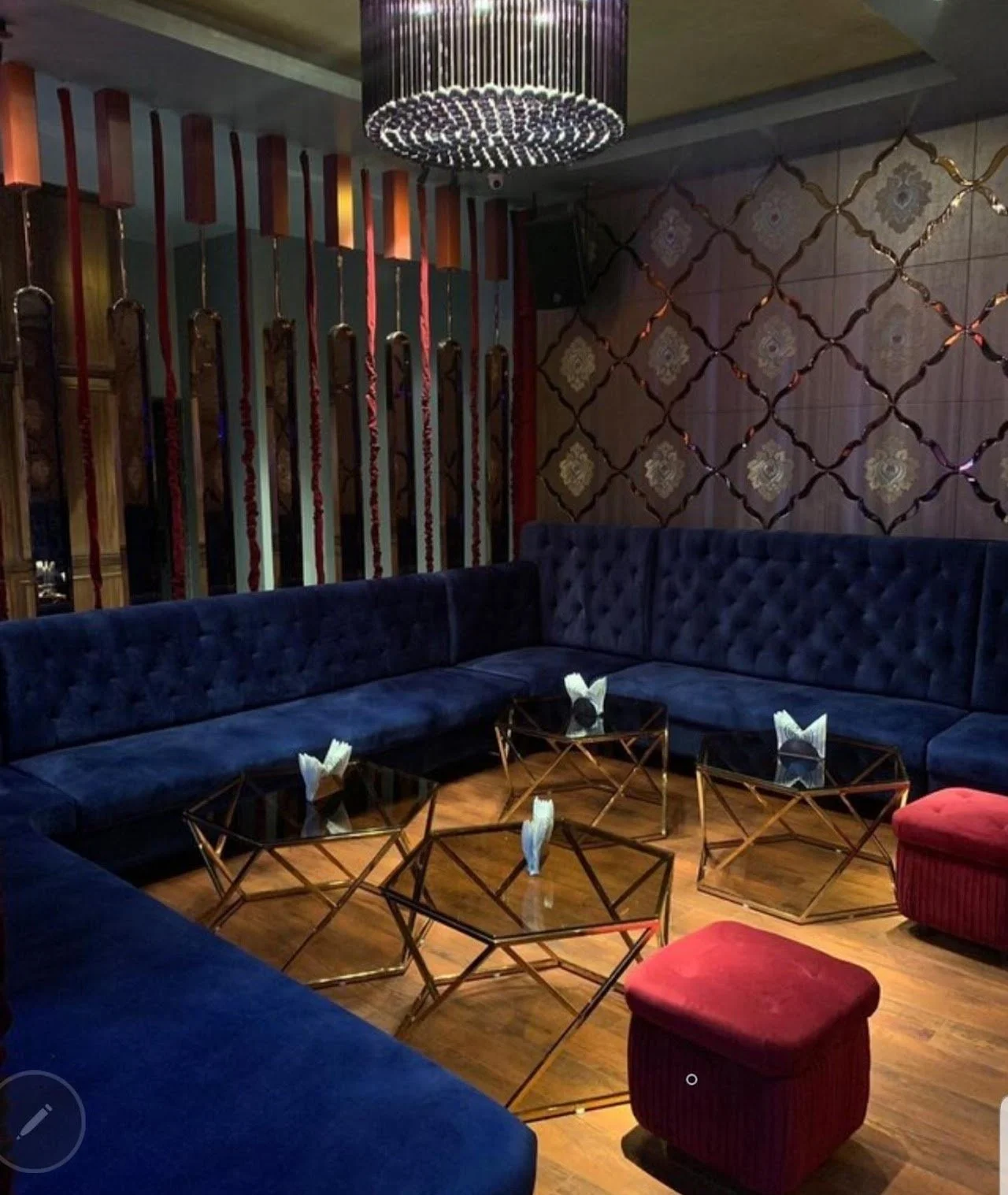 A luxurious lounge with dark blue tufted seating arranged in a U-shape, gold geometric coffee tables with napkins on top, a red velvet ottoman, a decorative wall with gold patterns, and vertical red and gold accents on a partition with a large chande