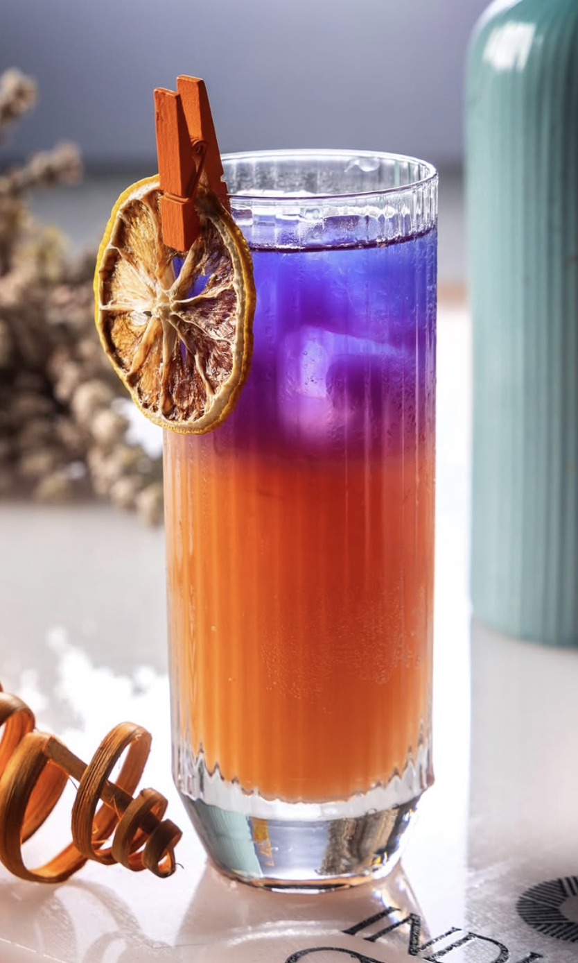 Colorful layered cocktail in a tall glass garnished with a dried lemon slice attached to a small orange clothespin.