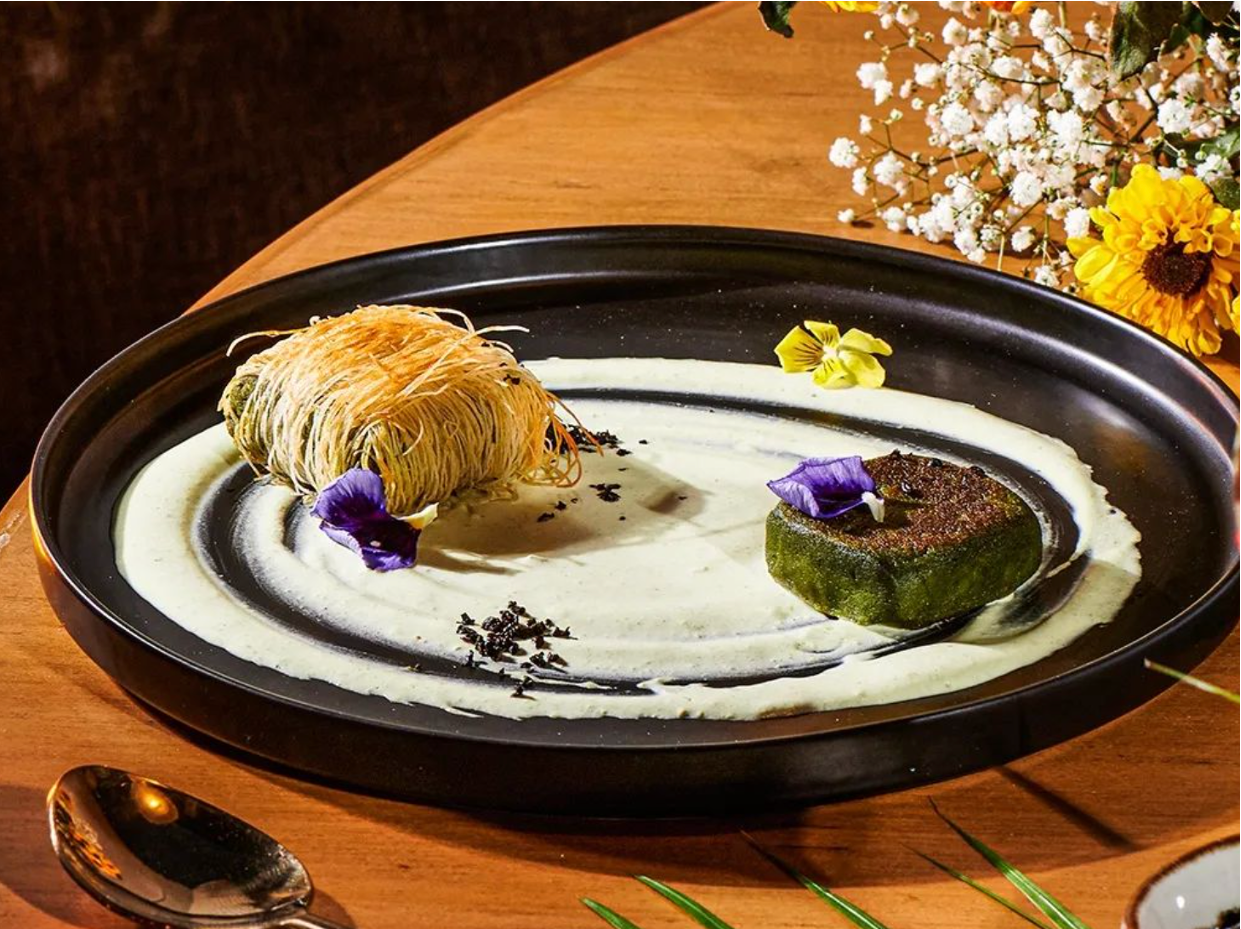 A black plate with a swirl of green sauce and two decorative flowers, one purple and one yellow, with a crispy nest-like food item and a rectangular green item garnished with flowers. There are flowers on a nearby table in the background.