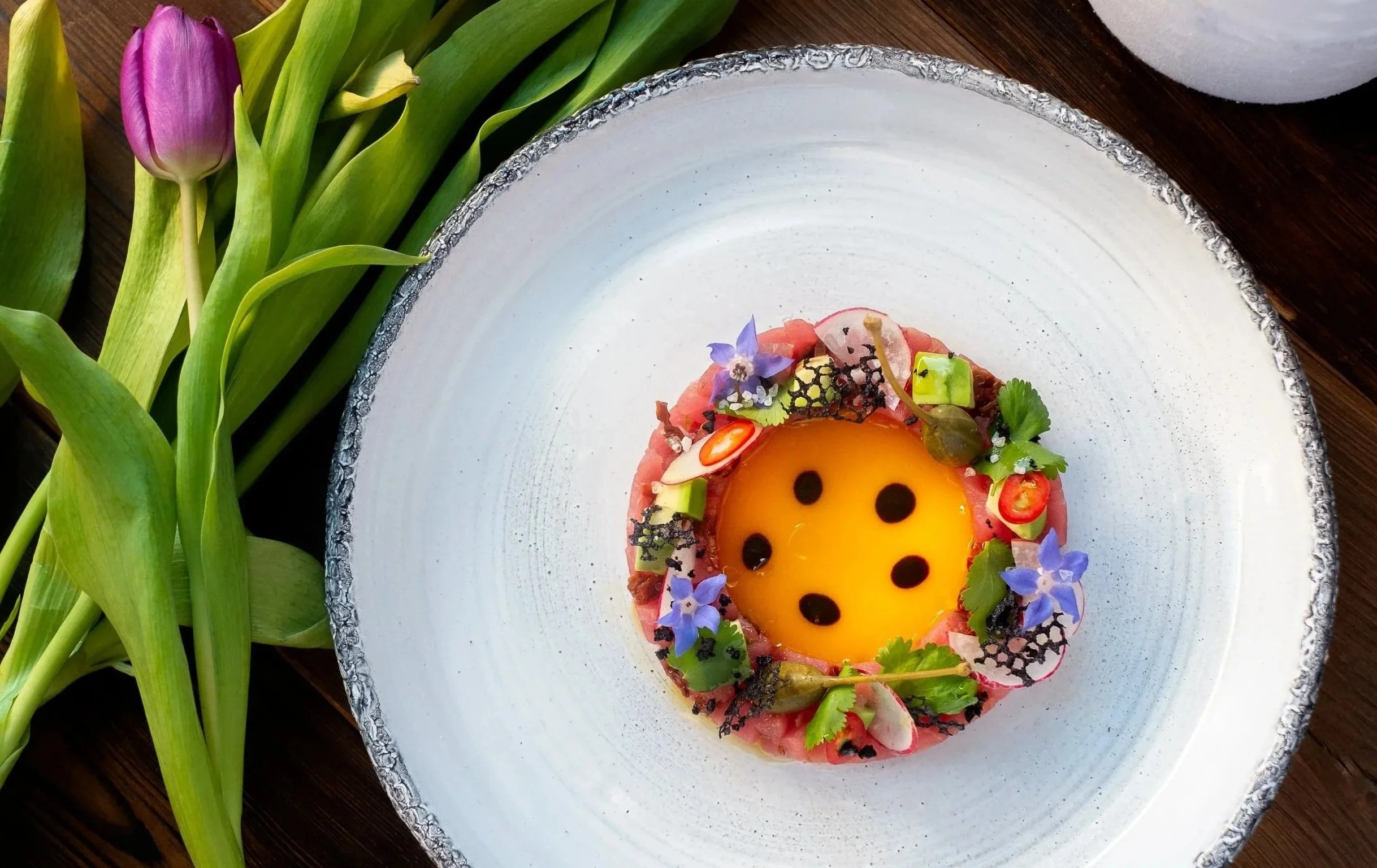 A decorative dish with a yolk-like center, topped with black dots, surrounded by various small vegetables, flowers, and garnishes, on a white plate with a textured rim, next to a bunch of purple tulips.