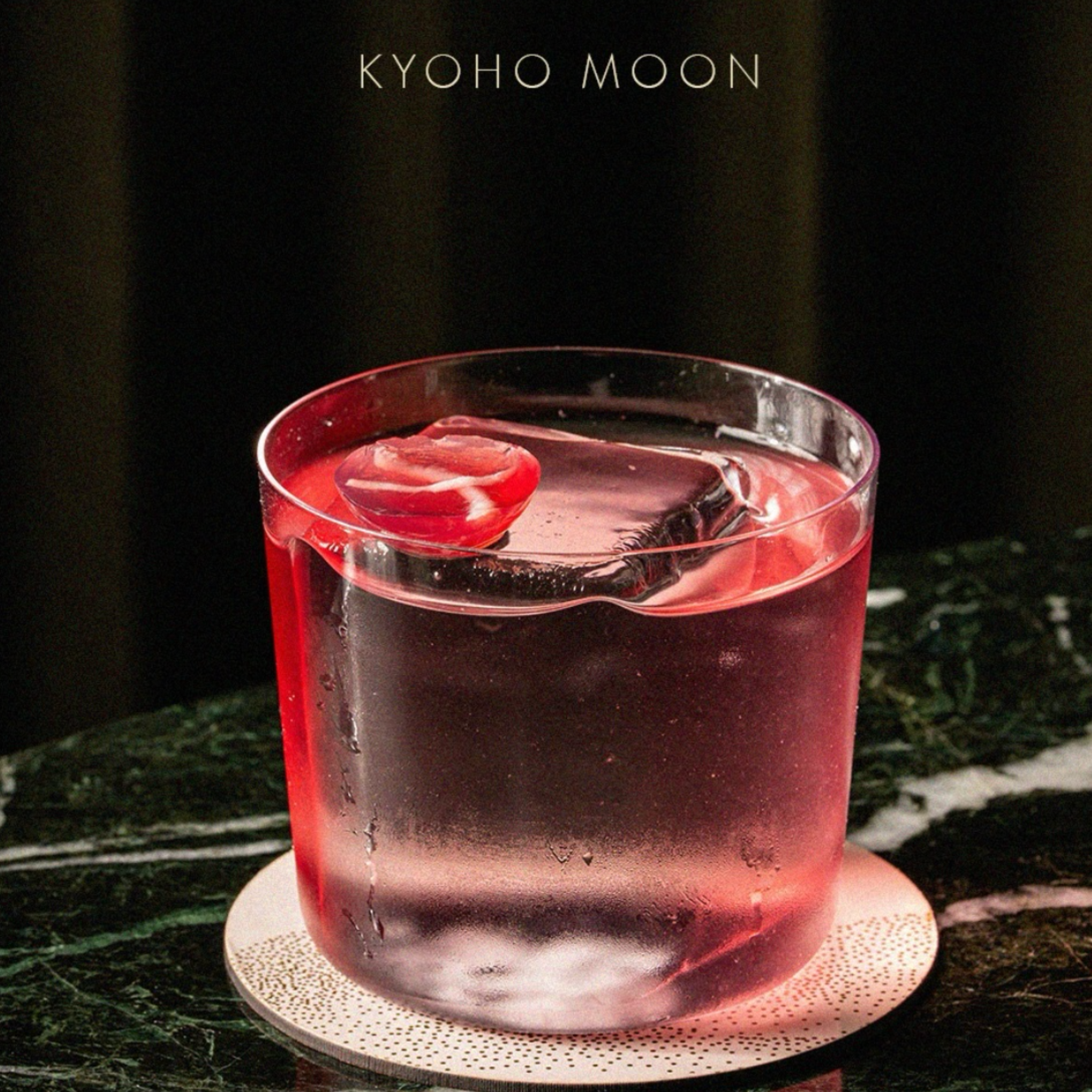 Close-up of a pinkish-red drink in a clear glass with ice and slices of fruit, placed on a patterned white coaster on a dark surface.