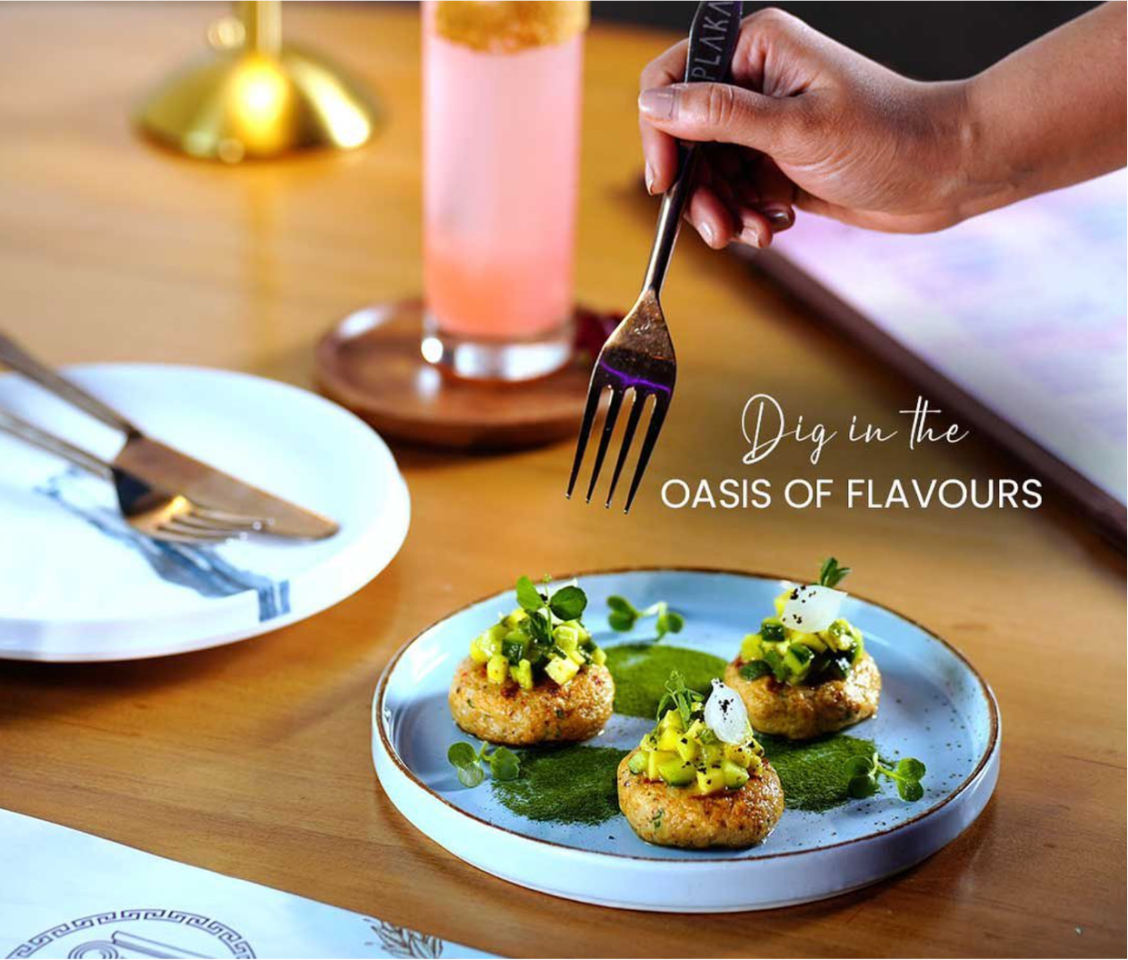A plate with three gourmet appetizer bites garnished with herbs and green vegetables. A hand with a fork hovers above them, holding a black utensil with the words 'PLAKAS' on it. Background includes a pink drink on a wooden coaster, a white plate wit
