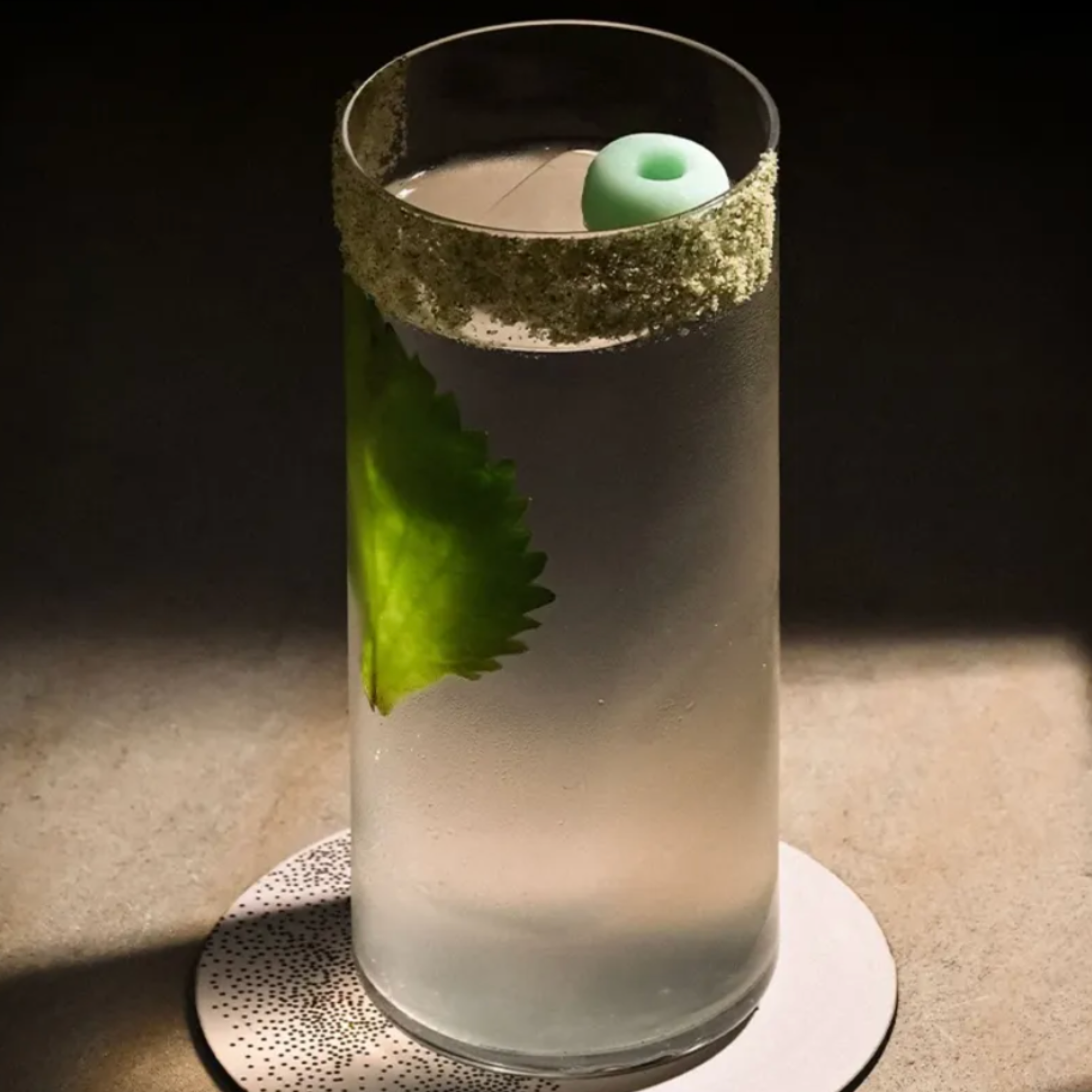 A white cylindrical container holding a green-colored die with a hollow center, surrounded by a narrow band of small white beads or crystals, set on a white, round plate with black dotted patterns, on a gray surface with a dark background.