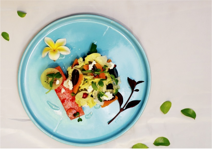 A plate of colorful salad with vegetables and edible flowers on a blue plate, decorated with green leaves and a yellow flower.