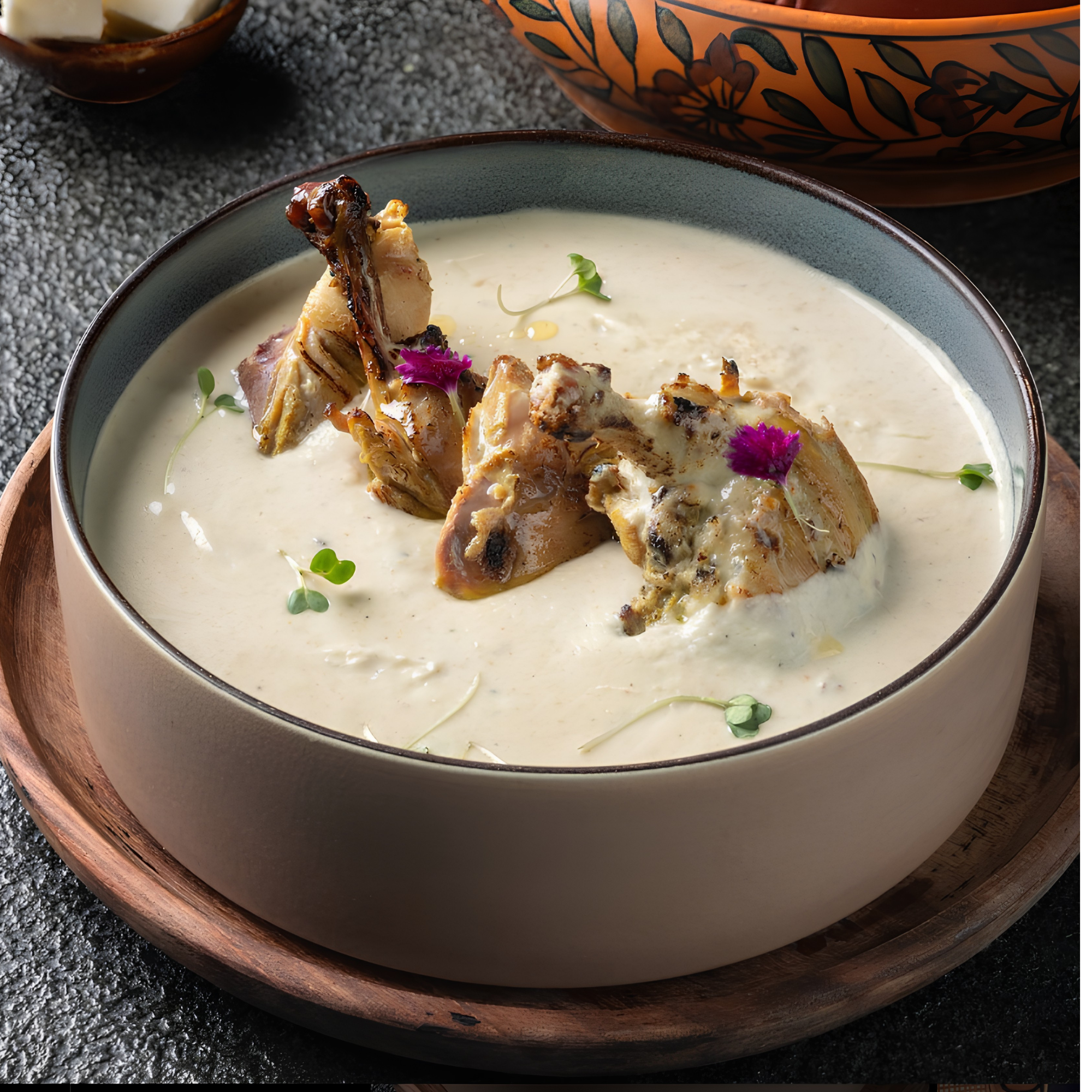 A bowl of creamy soup topped with grilled chicken drumsticks, edible flowers, and microgreens, served on a wooden tray with a decorative orange and black bowl in the background.
