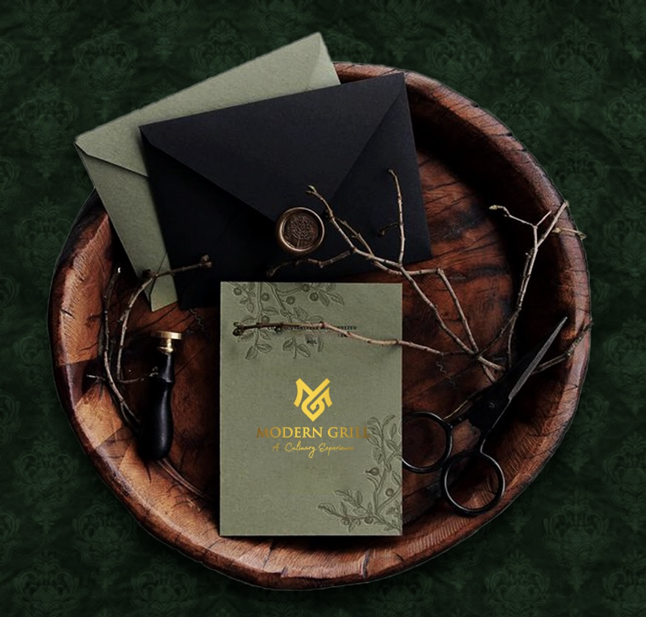 Wooden tray with two envelopes, a business card with 'Modern Grill' logo, a branch, scissors, and a wax seal on dark green patterned background.