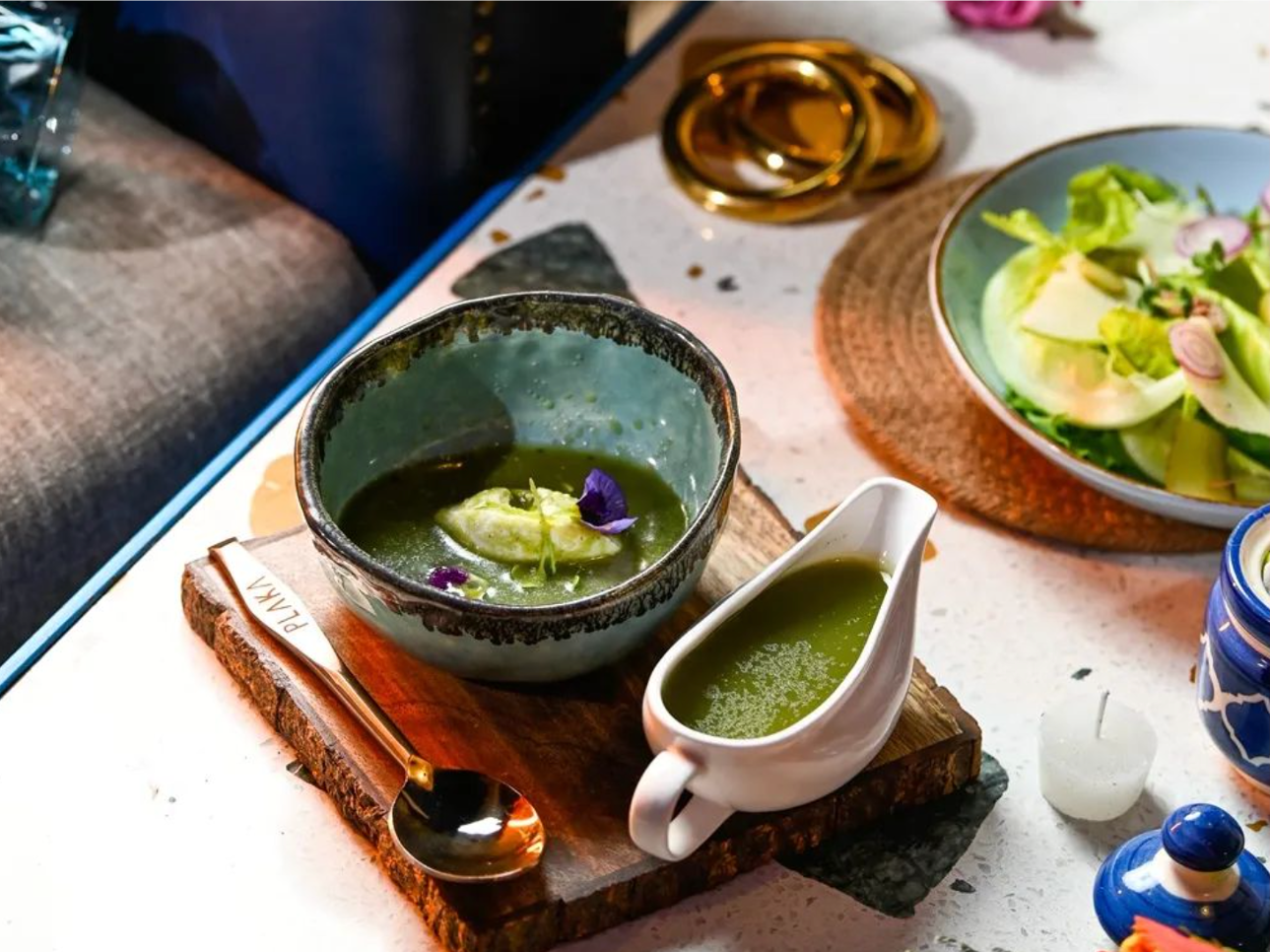 A ceramic bowl and a white creamer filled with green matcha tea, garnished with edible flowers, sit on a wooden board. To the right, a salad with greens and sliced vegetables is in a bowl, and a small white plant candle is nearby on a table.