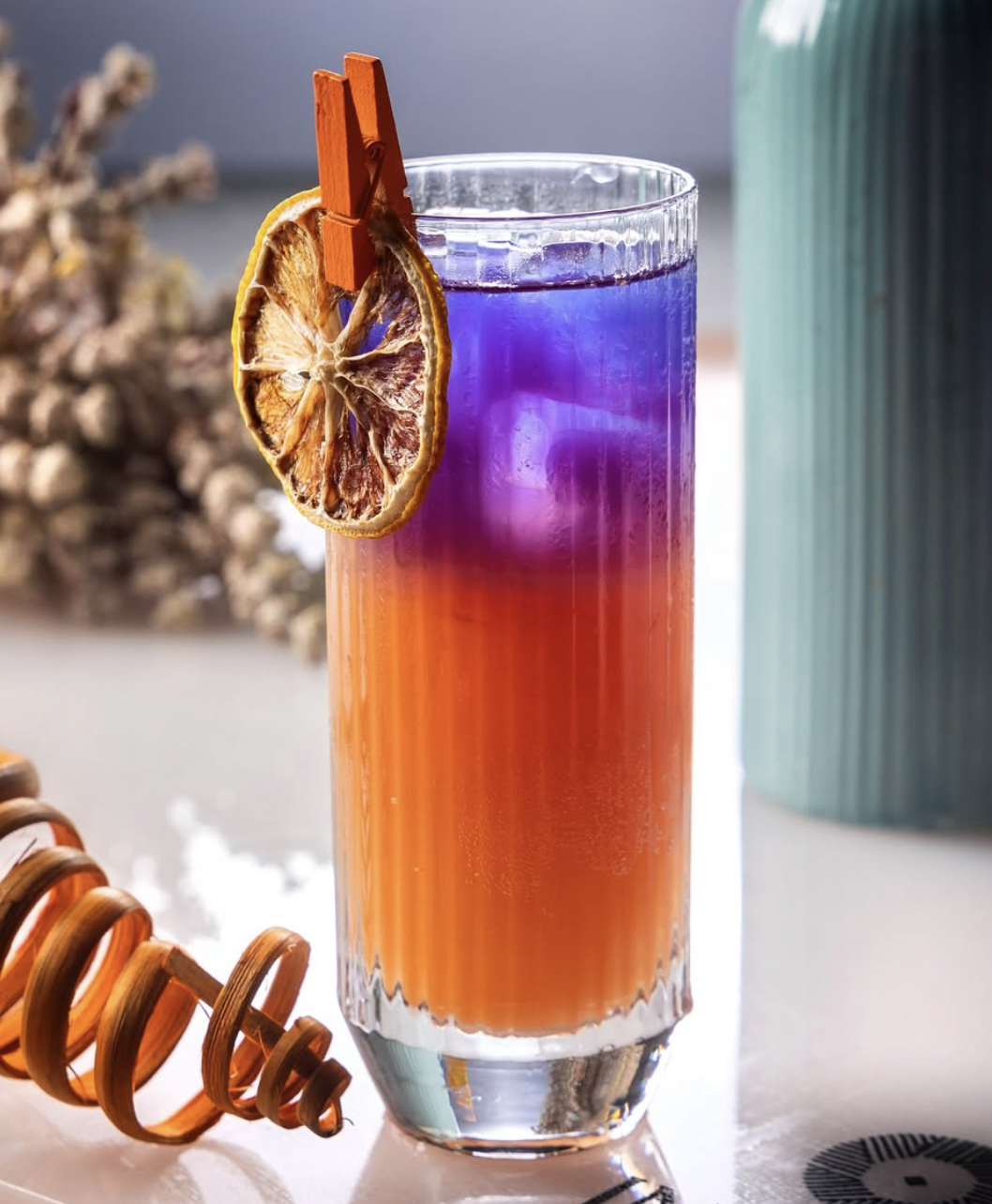 Colorful layered cocktail in a tall glass garnished with a dried lemon slice and small wooden clothespin, with a background of blurred objects and a light-colored surface.