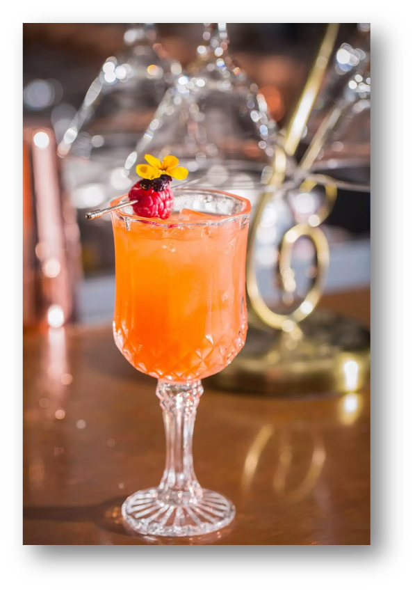 An elegant glass filled with a pink cocktail, garnished with a raspberry and a small yellow flower, placed on a wooden surface with barware in the blurred background.