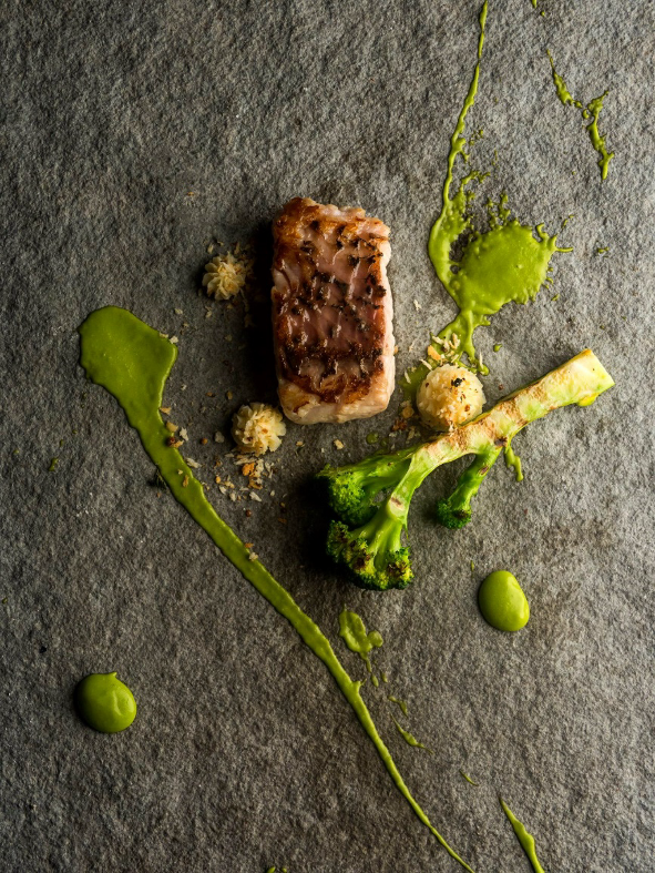 A plated dish featuring a piece of seared fish with a brown crust, accompanied by broccoli, cauliflower, and green purée, with artistic green sauce splatters on a dark textured surface.