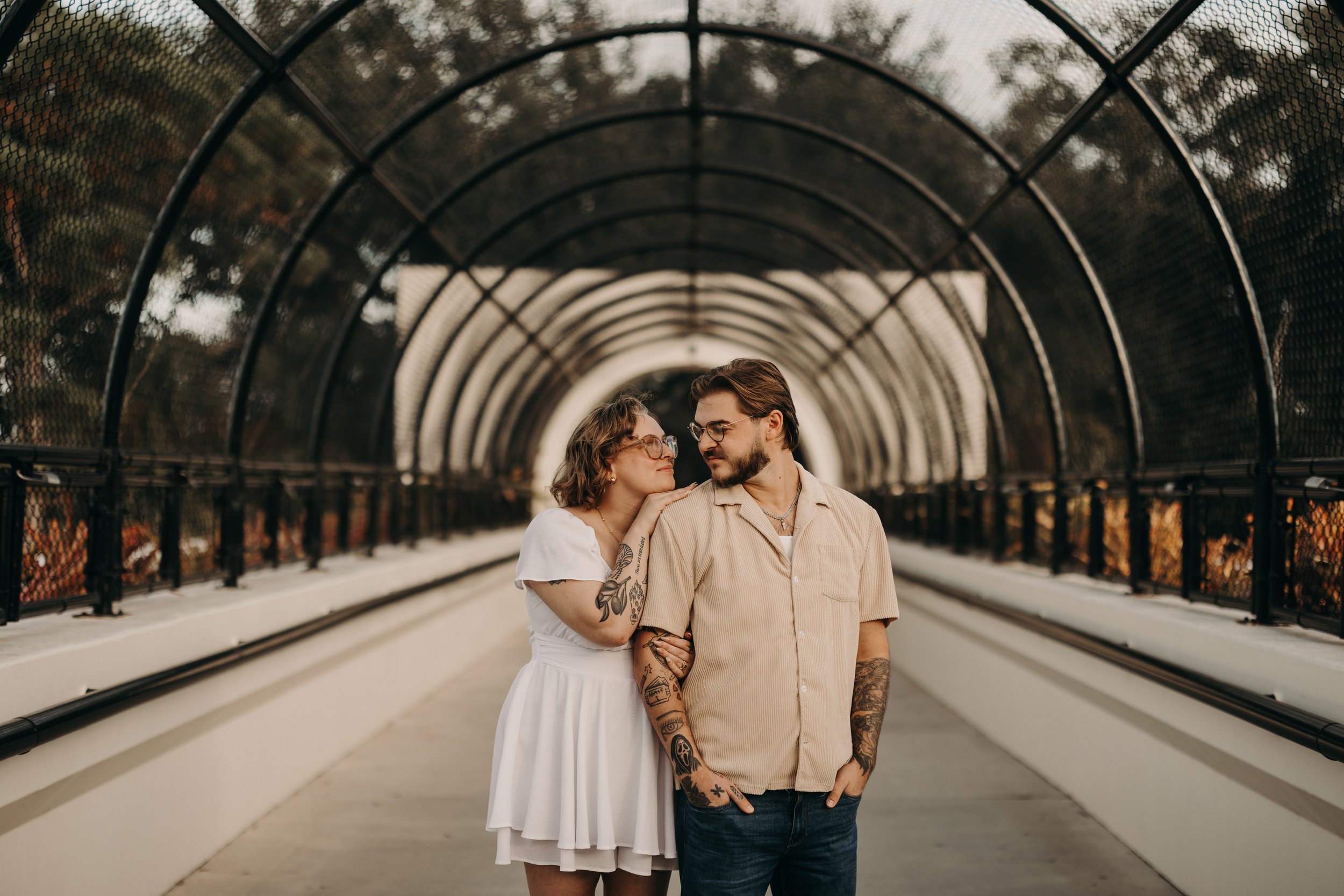 A couple stands close together under a tunnel, looking into each other's eyes. The woman wears a white dress and glasses, has tattoos on her arms, and has her hand on the man's shoulder. The man wears a beige shirt, has tattoos on his arms, and wears glasses.