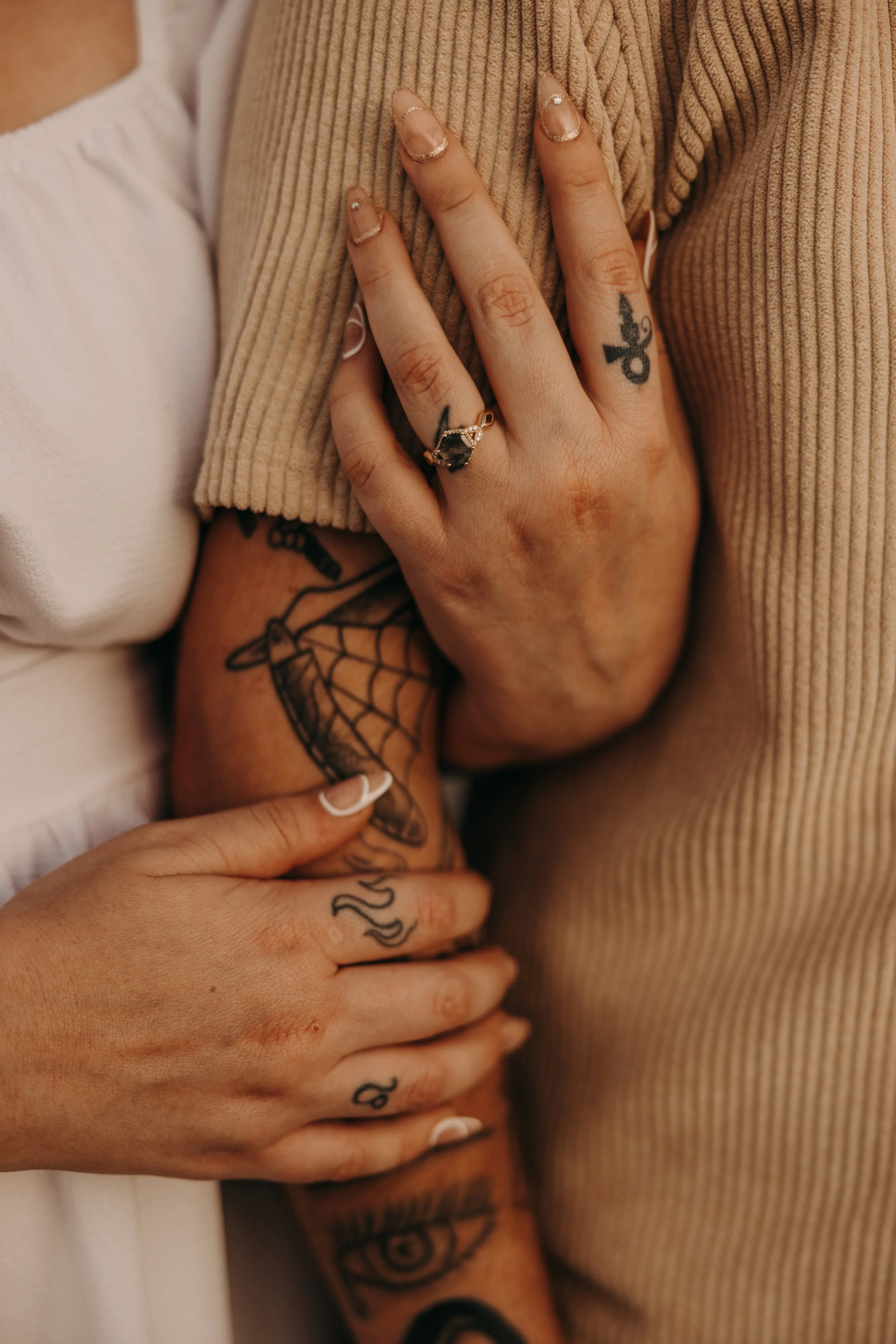 Close-up of two people's hands intertwined, showing tattoos and a ring, with clothing in beige and white tones.