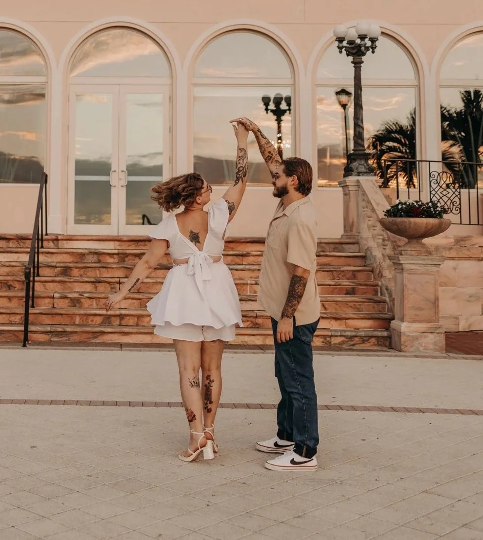 A couple dancing in front of a building with large arched windows during sunset, with palm trees in the background.