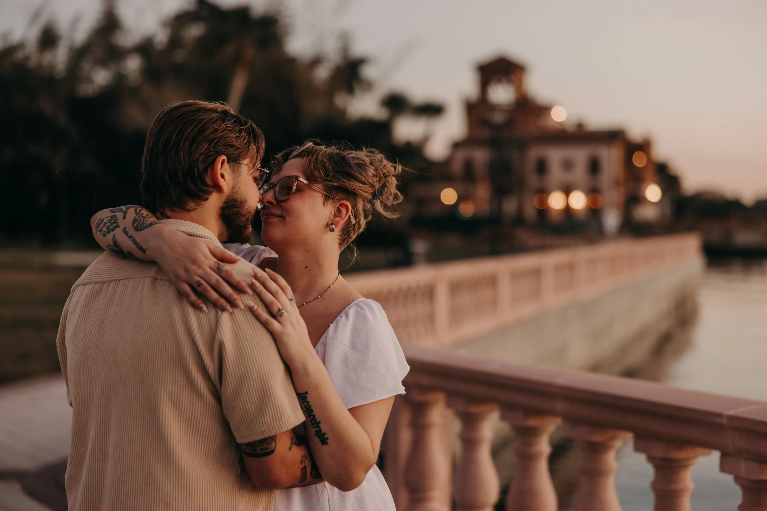 A couple embraces near a river at sunset, with a historic building and trees in the background.