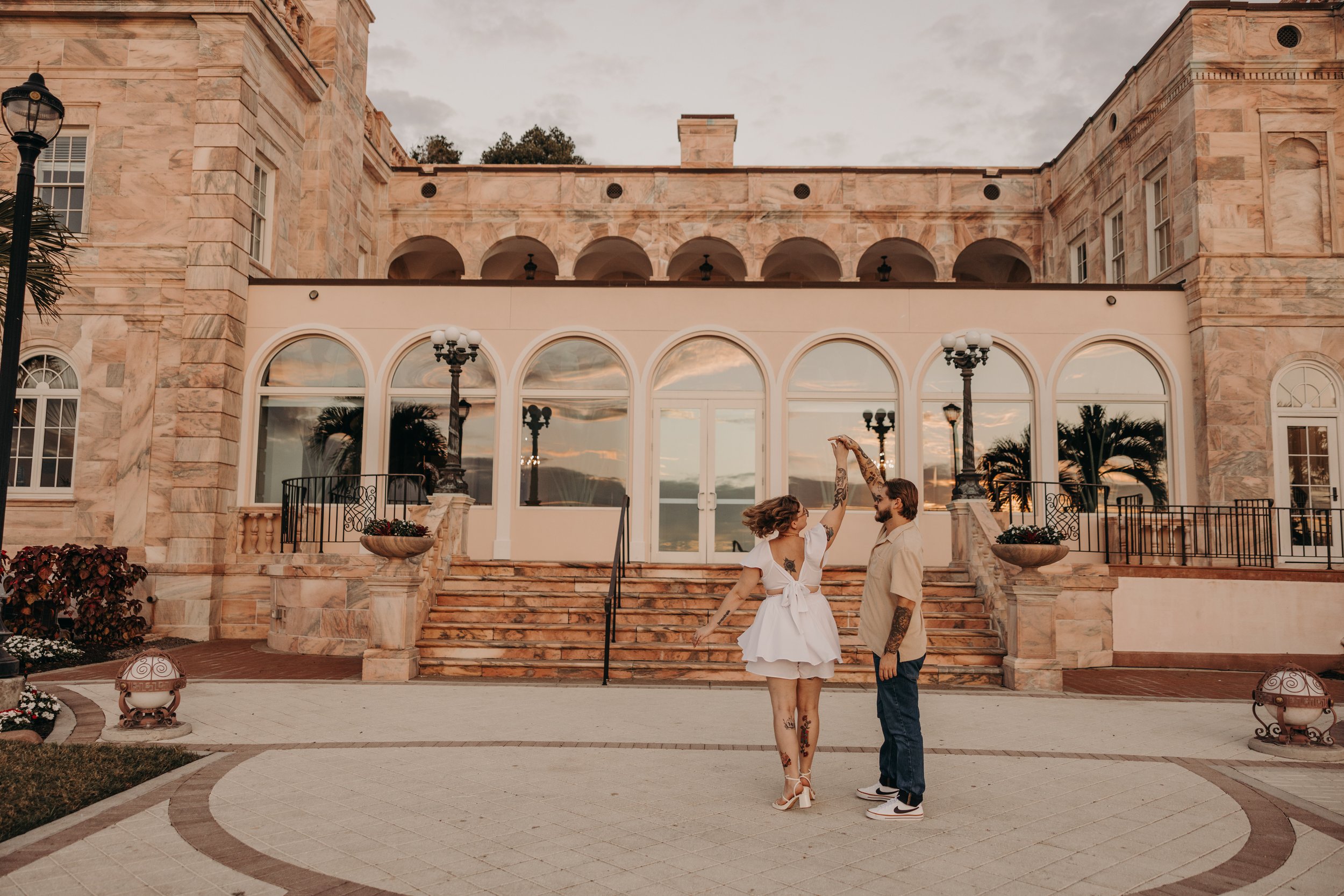 A couple dancing on the steps of a large mansion with arched windows and stone exterior at sunset.