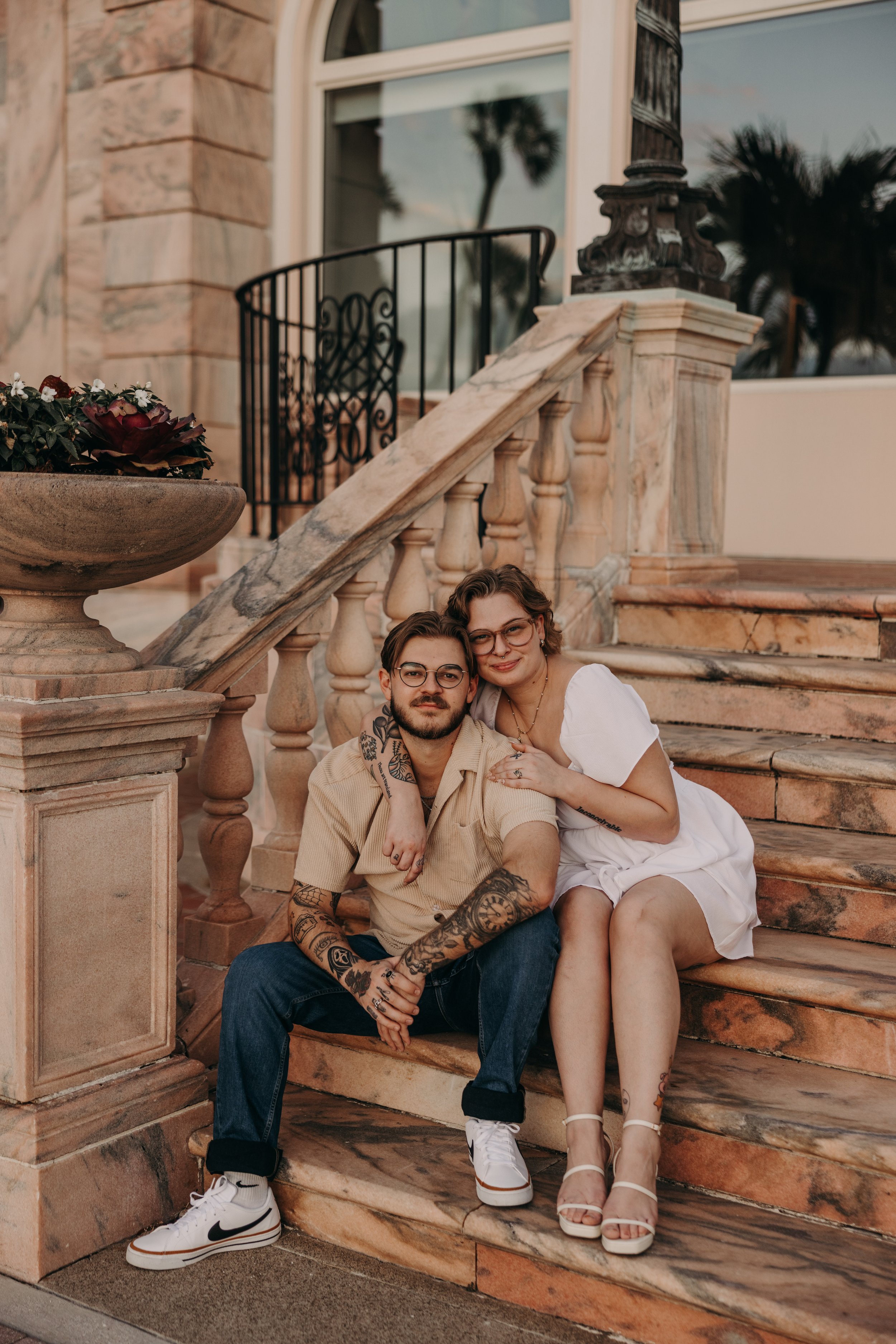 A couple sitting on marble stairs outside a building with palm trees reflected in the window behind them. The man has tattoos and glasses, wearing a beige shirt and dark jeans. The woman has glasses, tattoos, and curly hair, wearing a white dress and heels.