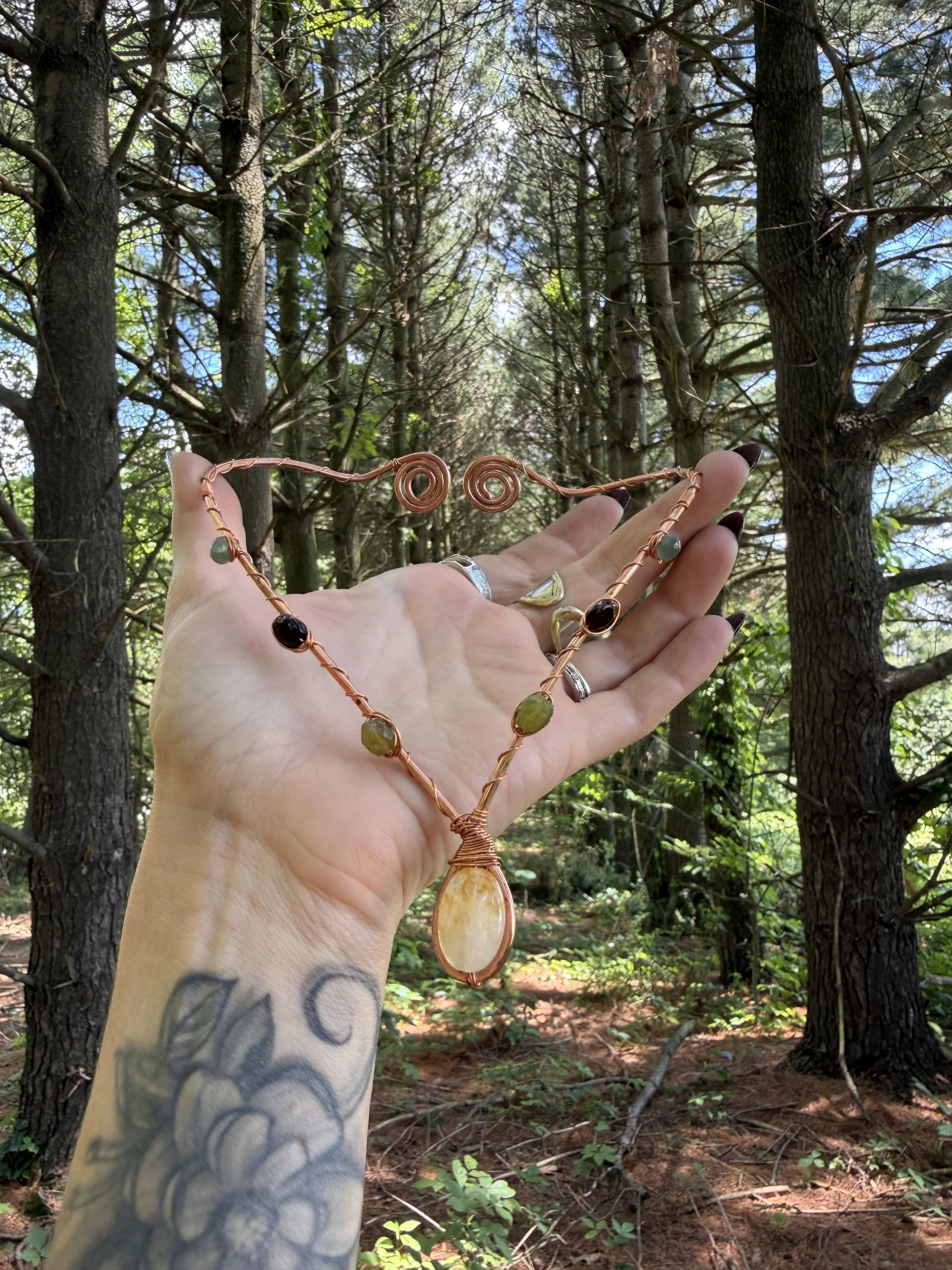 A hand with tattoos holding a large copper wire wrapped necklace with golden healer quartz, emerald, vesuvianite, and garnet stones and a large pendant, raised against a background of tall trees in a forest.