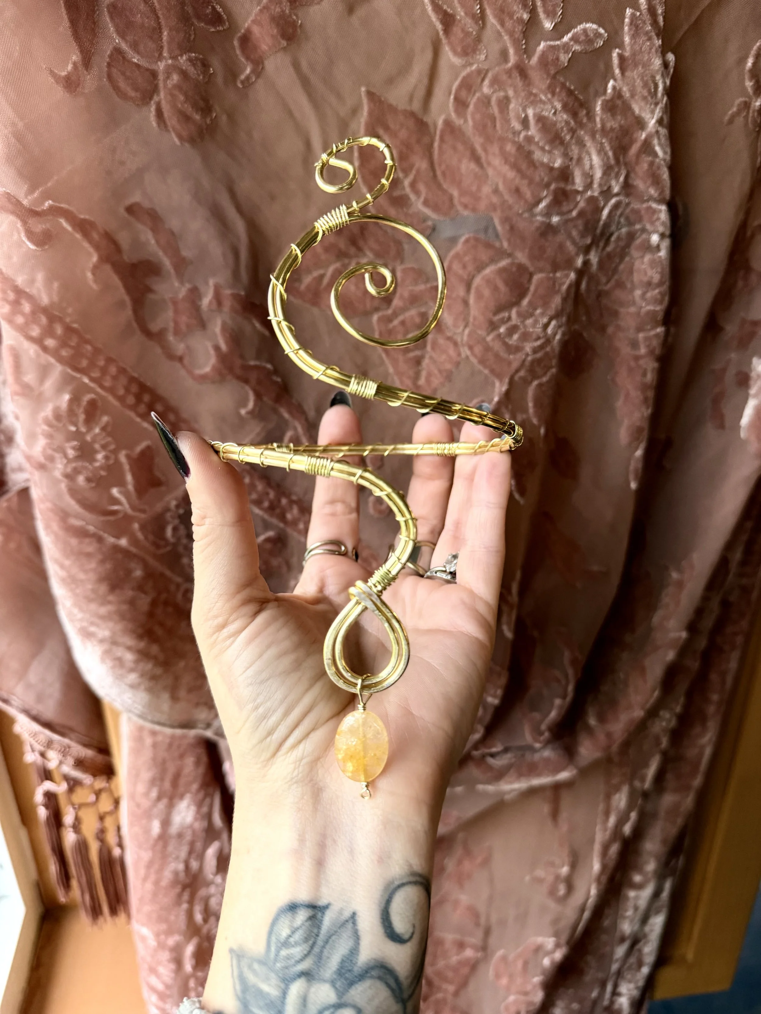 A person holding a gold wire spiral upper arm cuff with a dangling golden healer quartz stone against a background of pink, embroidered fabric.