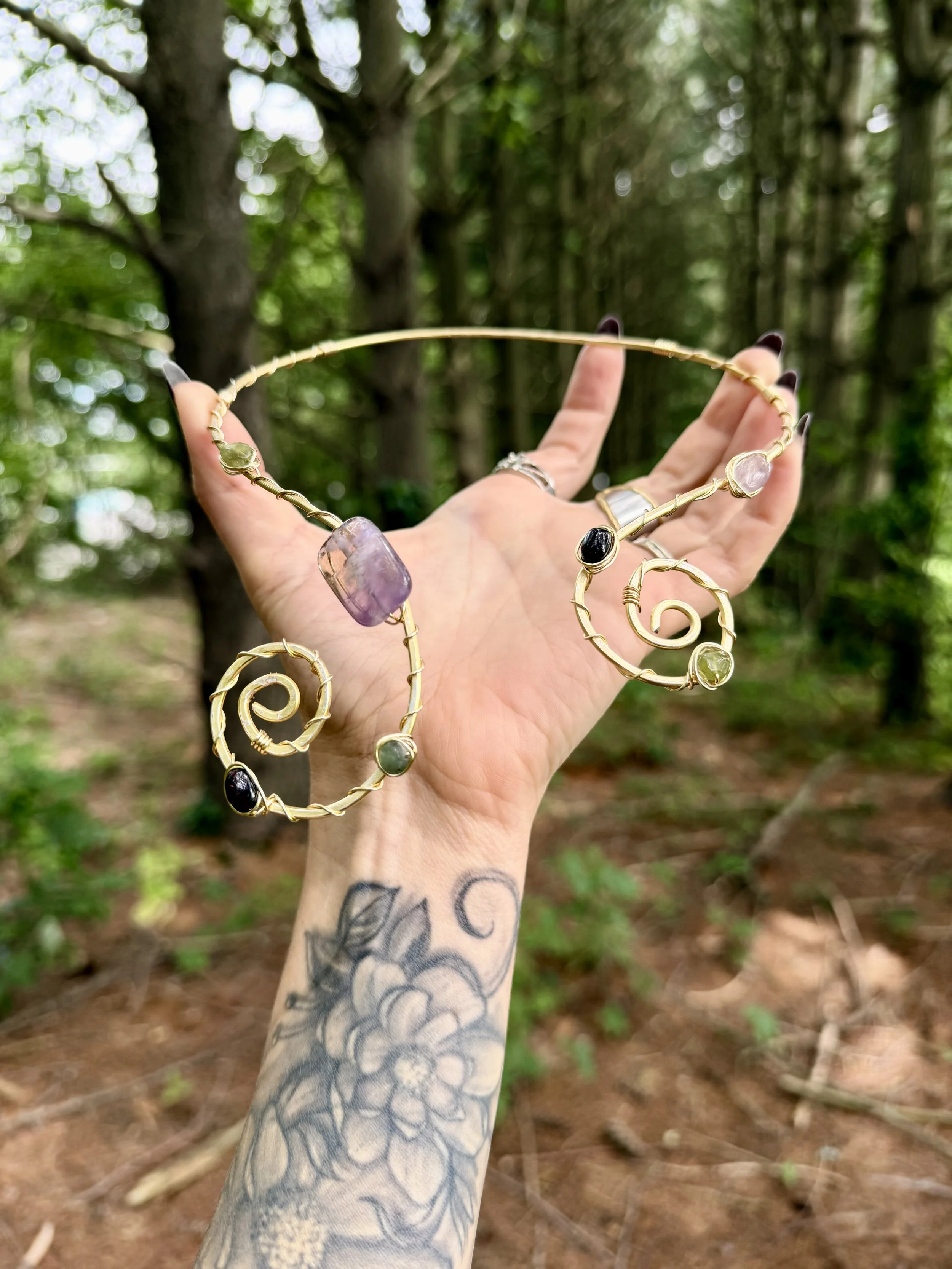 A hand with a tattoo holding a gold jewelry necklace with large colorful amethyst, garnet, vesuvianite, emerald gemstones outside in a wooded area.